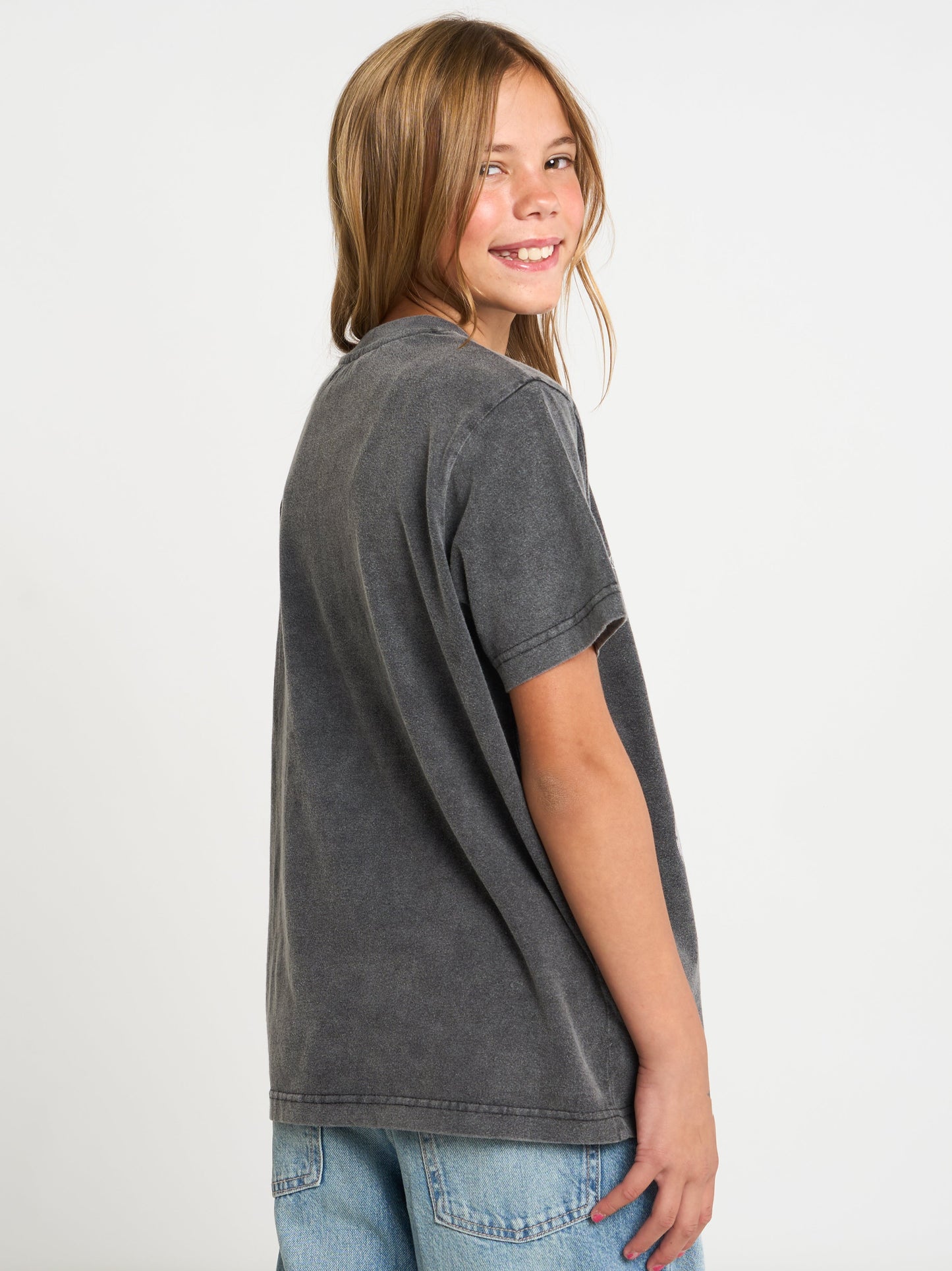 A young girl with light brown hair is shown from a side-back angle wearing a washed dark gray short-sleeve t-shirt and light blue jeans against a plain white studio background.