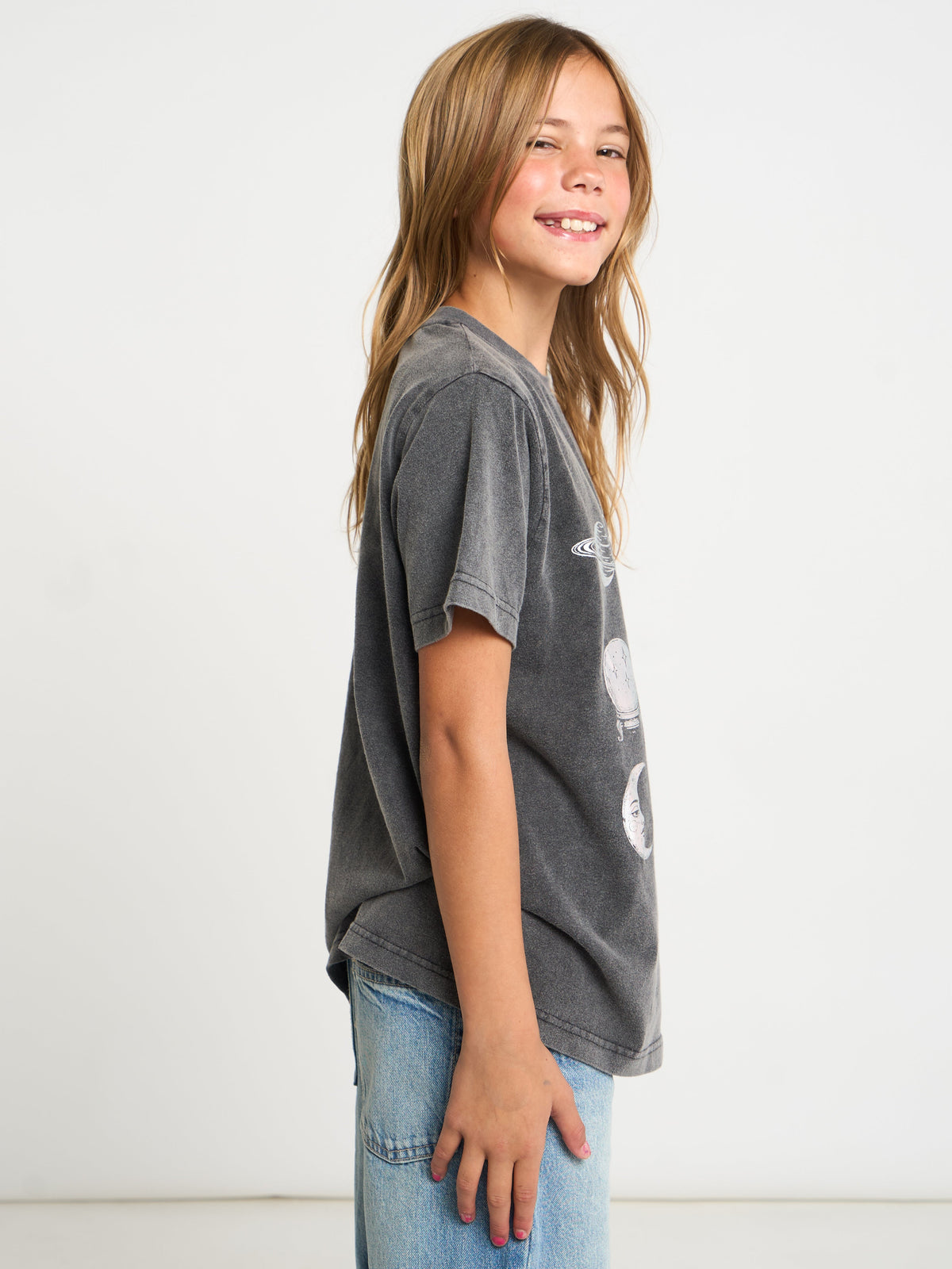 A young girl with long light brown hair is shown in profile wearing a washed charcoal gray graphic t-shirt and light blue jeans against a plain white studio background.