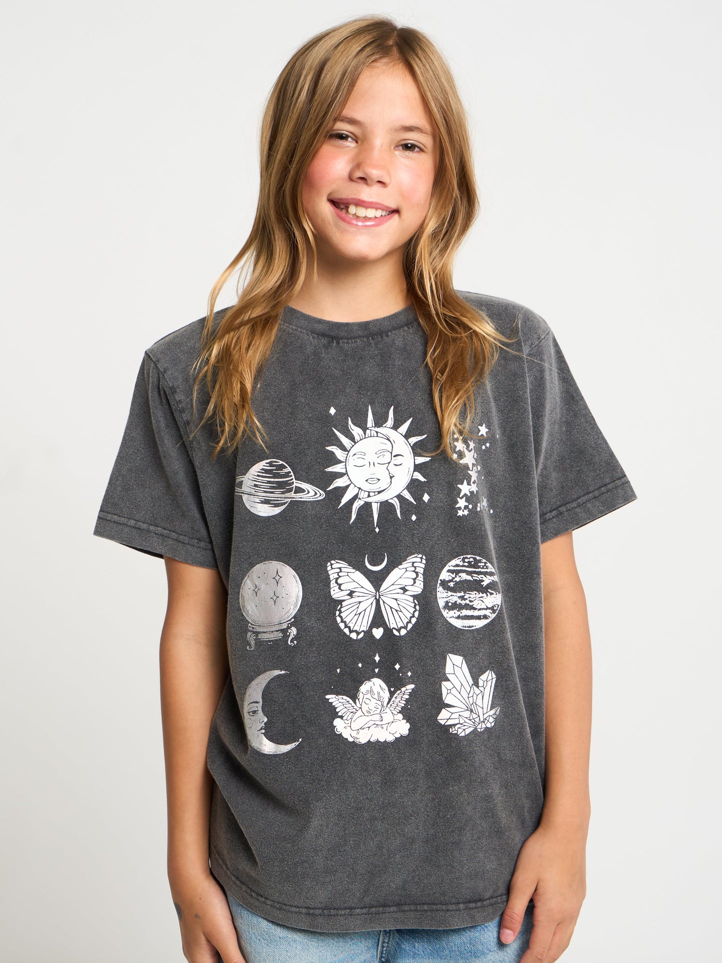 A smiling young girl with blonde hair wearing a dark gray mineral-wash graphic t-shirt with white celestial illustrations, paired with light blue jeans, against a light gray studio background.
