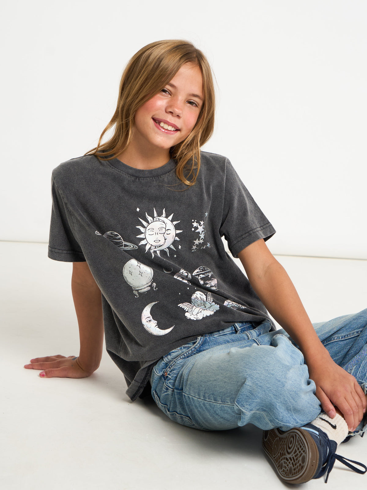 A smiling girl with blonde hair sits on a white floor wearing a washed black graphic t-shirt with celestial designs, blue jeans, and sneakers.