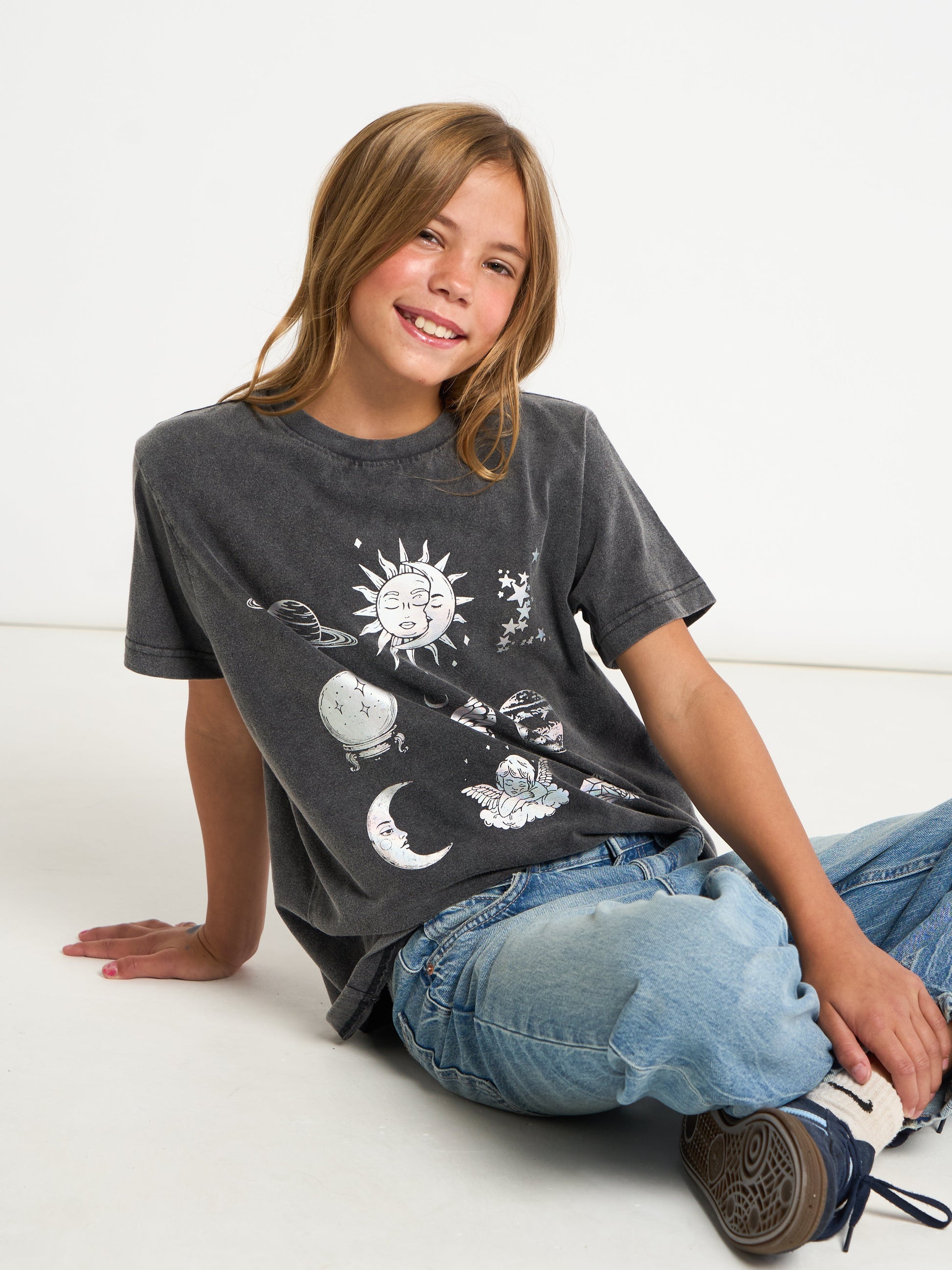 A smiling girl with blonde hair sits on a white floor wearing a washed black graphic t-shirt with celestial designs, blue jeans, and sneakers.
