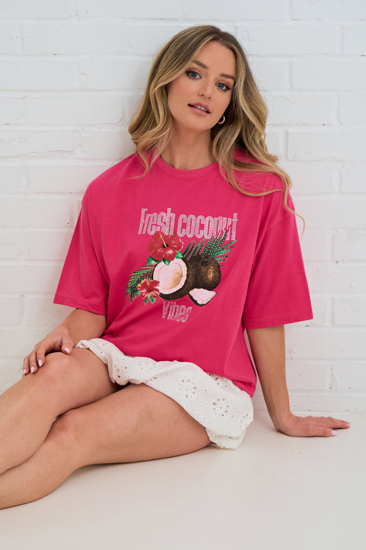 Woman wearing a pink t-shirt with a coconut graphic design against a white brick wall.