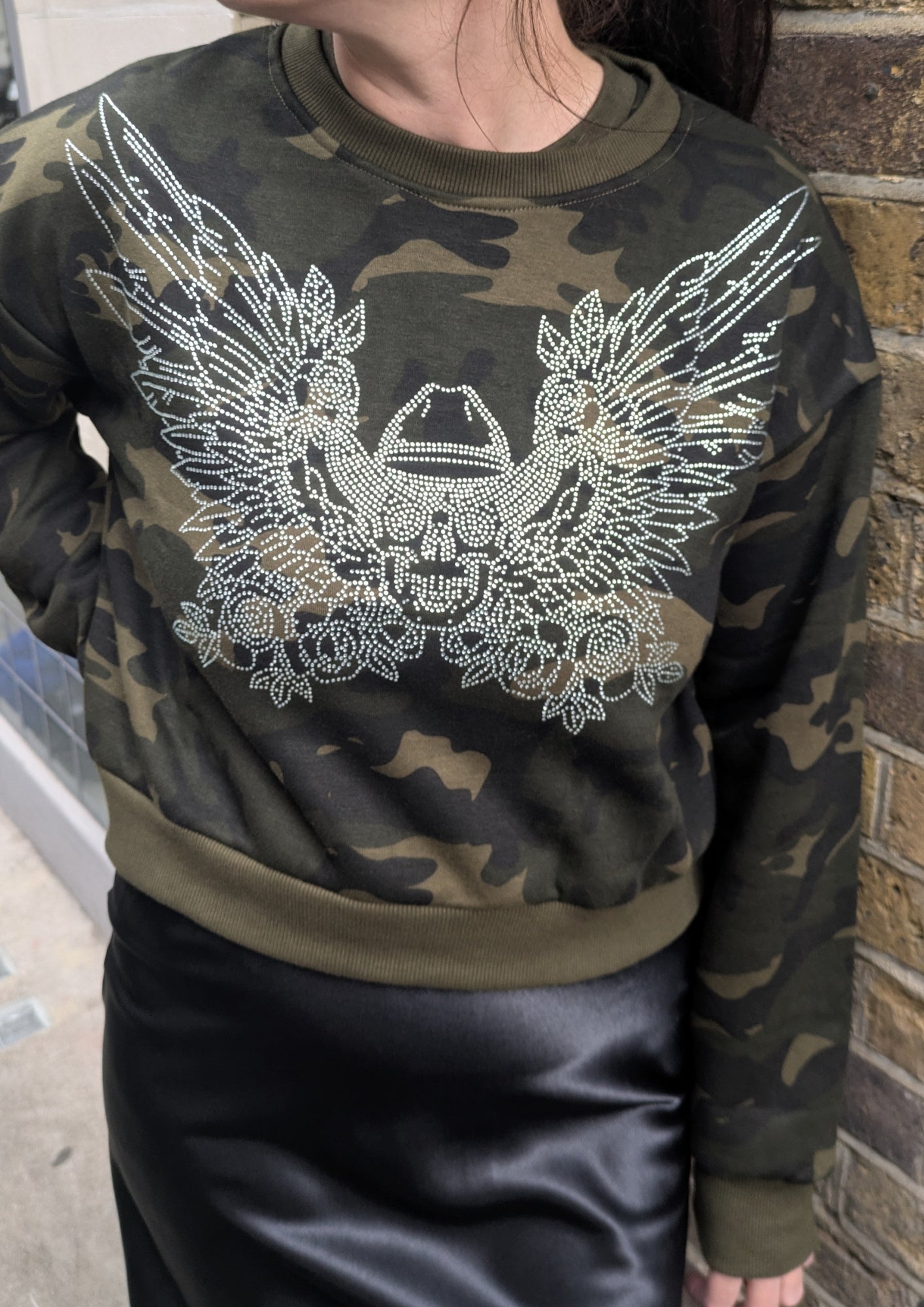Camouflage sweatshirt with a decorative rhinestone work of a cowboy skull with angel wings worn by a person against a brick wall.