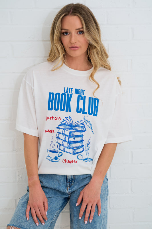 Person wearing a 'Late Night Book Club' t-shirt with a graphic design against a white brick wall.