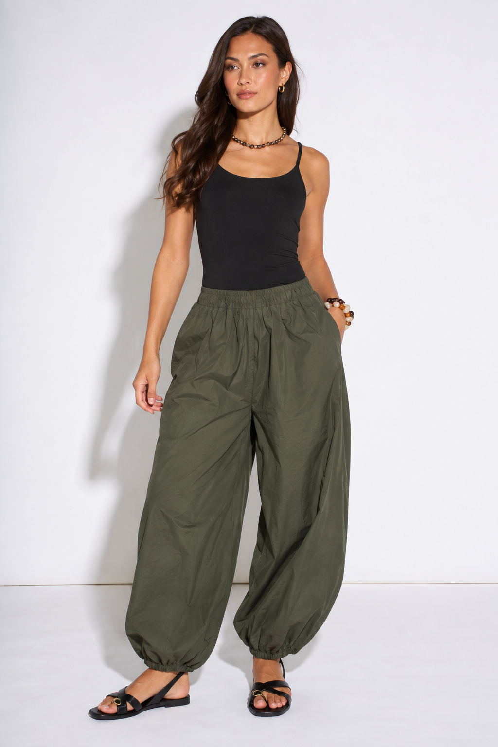 A woman modeling a pair of loose, wide-leg olive green pants with elastic cuffs, paired with a black tank top and black flat sandals, standing against a white studio background.