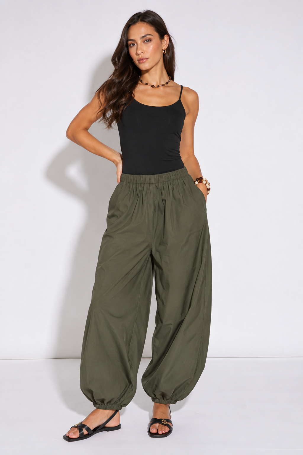 A woman modeling a pair of loose, wide-leg olive green pants with an elastic waist and gathered cuffs, paired with a black tank top and black sandals, against a plain white studio background.