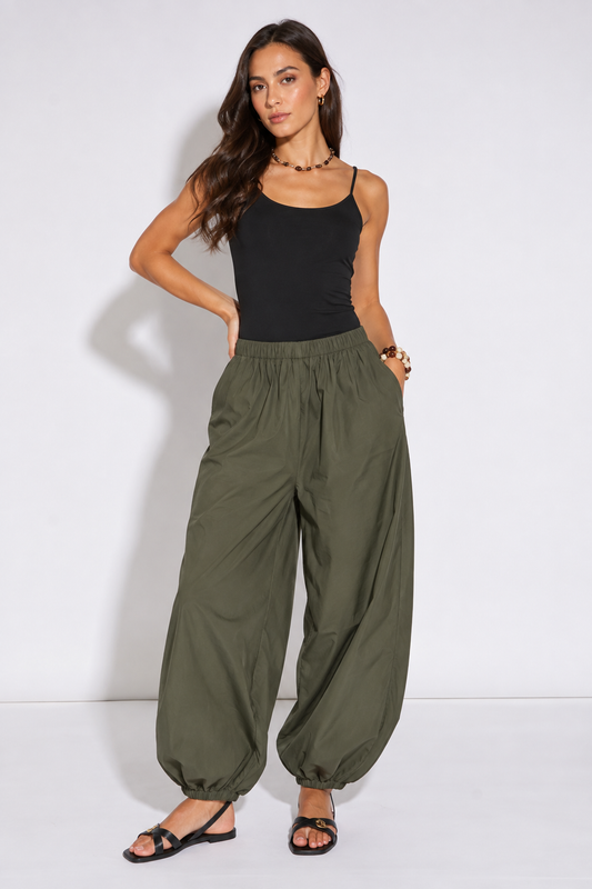 A woman modeling a pair of loose, wide-leg olive green pants with an elastic waist and gathered cuffs, paired with a black tank top and black sandals, against a plain white studio background.