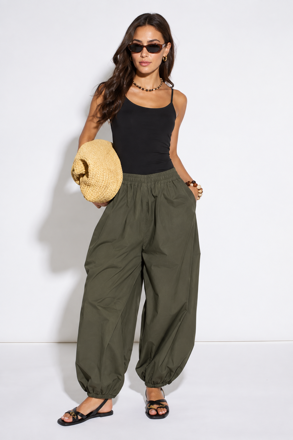 A woman modeling a pair of loose-fitting olive green harem-style pants with a black tank top, black sunglasses, and black sandals, holding a round straw hat against a plain white studio background.