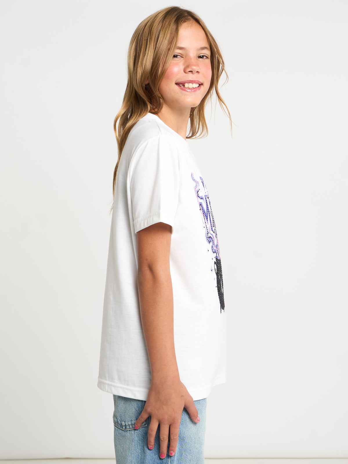 A young girl with blonde hair smiling while wearing a white short-sleeve t-shirt with a purple and black graphic print on the side, paired with light blue jeans, against a plain white background.