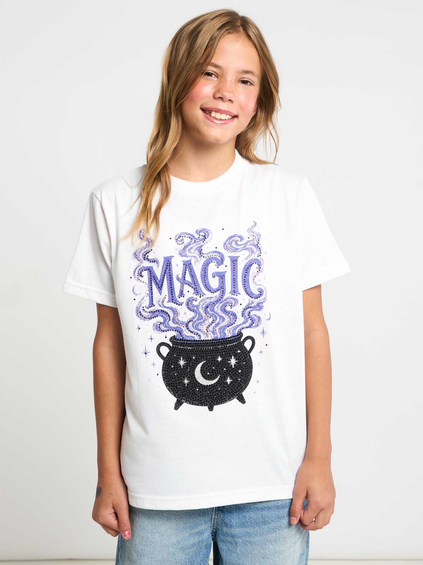 A young girl with blonde hair smiling while wearing a white graphic t-shirt with a purple and black 'MAGIC' cauldron design, paired with blue jeans, against a plain white background.