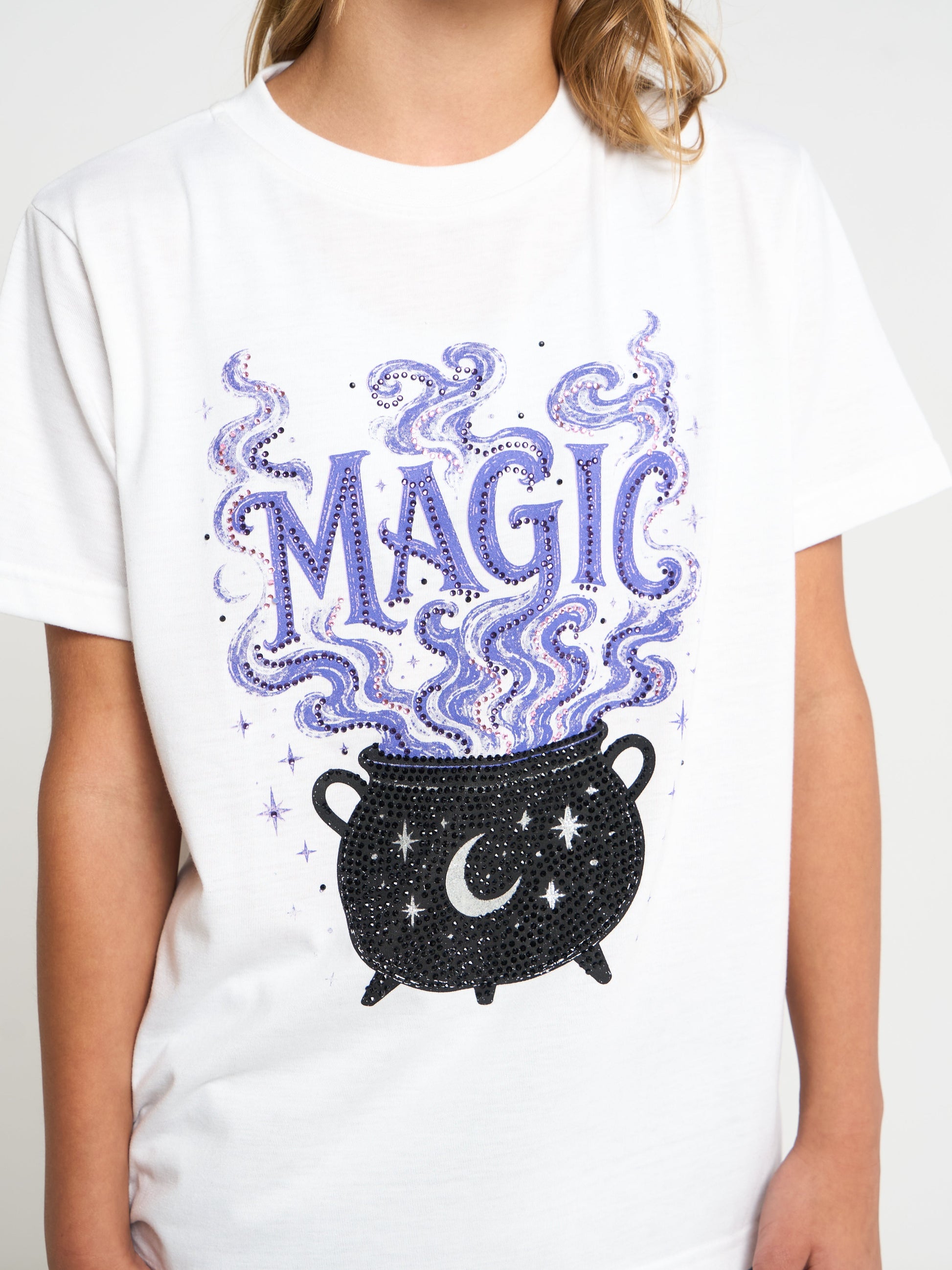 A white short-sleeve T-shirt worn by a person, featuring a large front graphic with the word "MAGIC" in purple outlined letters above a black cauldron with stars and a crescent moon, with purple smoke rising.