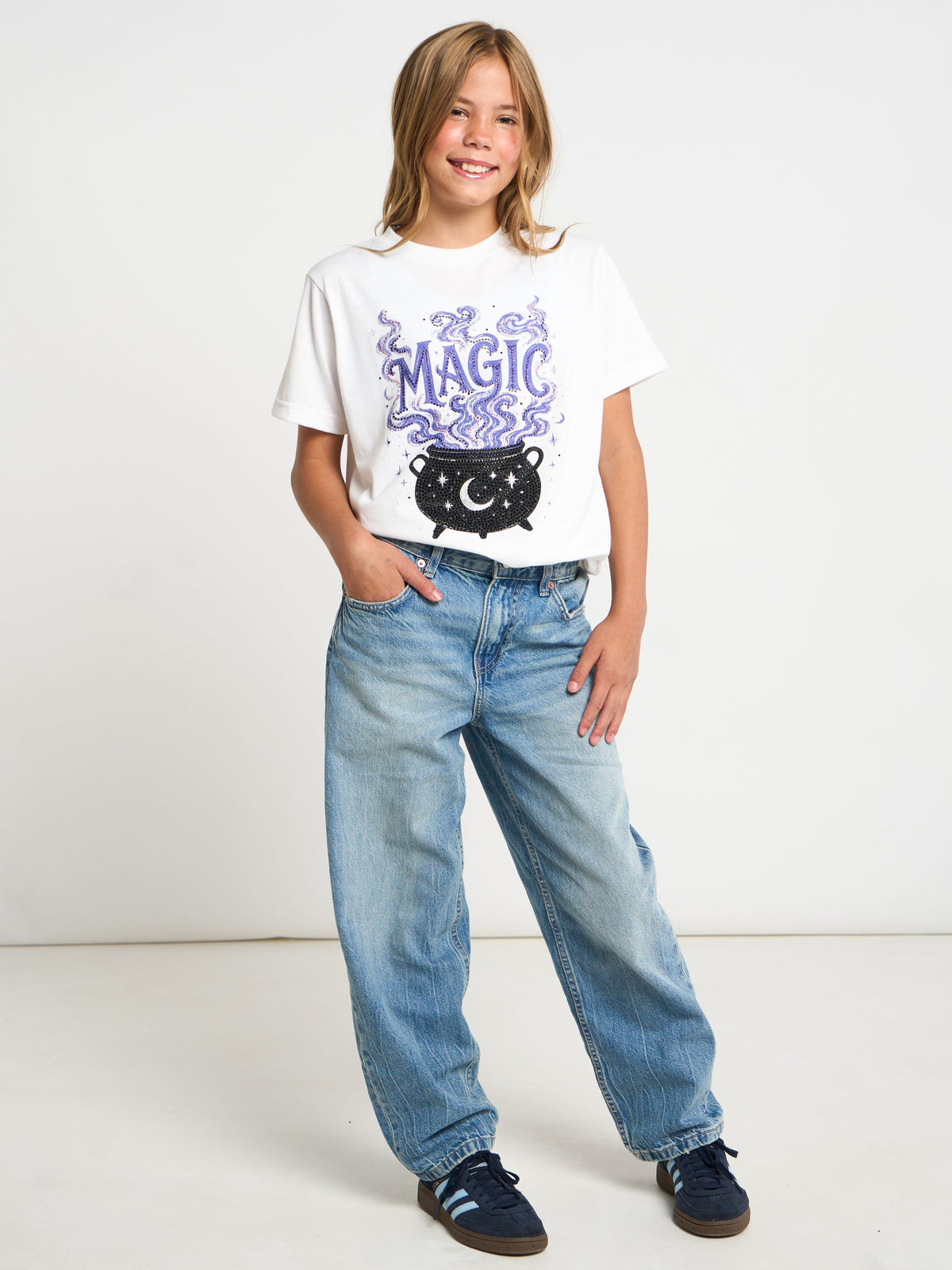 A child model wearing a white graphic t-shirt with a purple and black cauldron design, paired with light-wash blue jeans and dark blue sneakers, standing against a plain white studio background.
