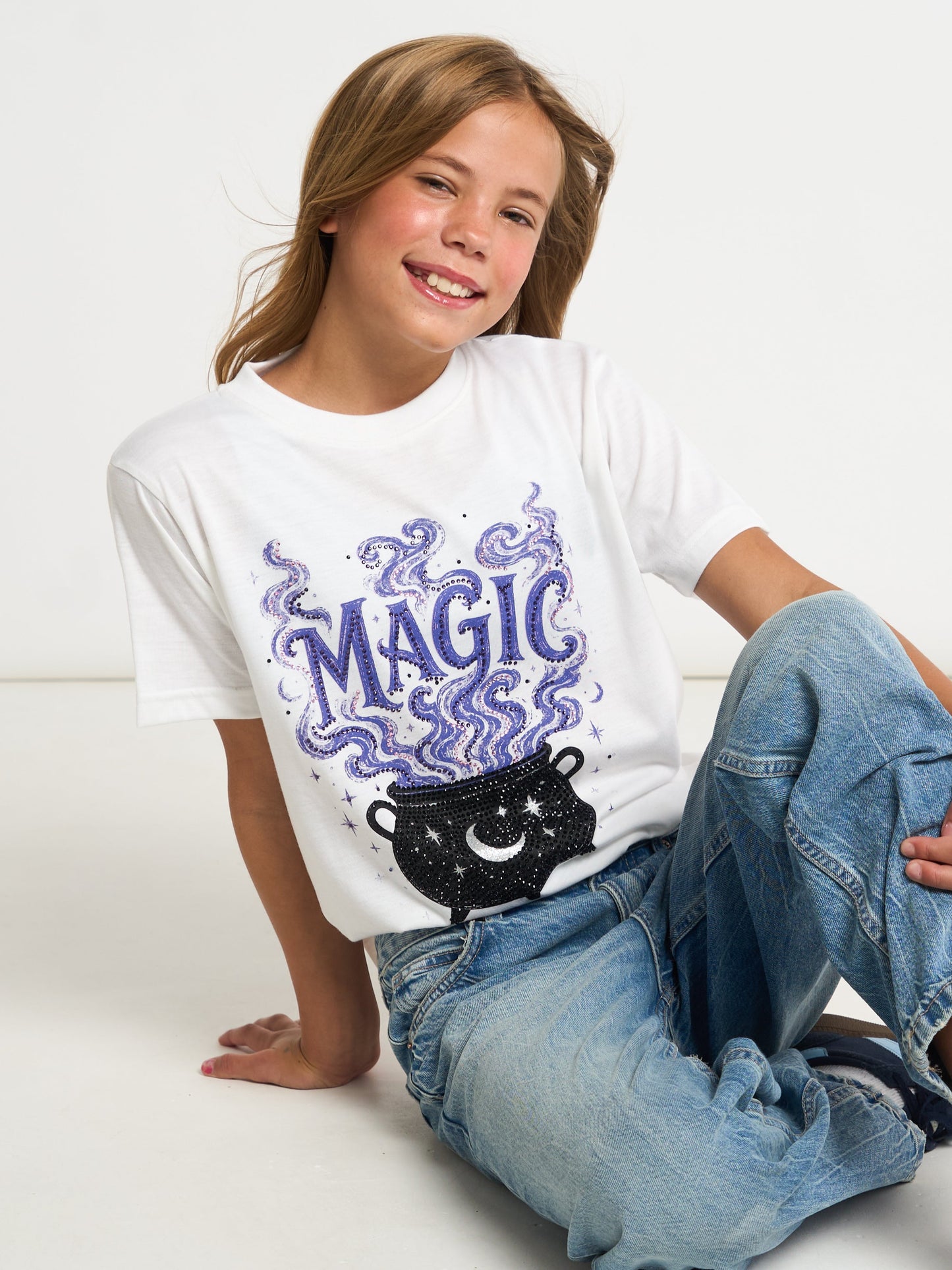 A smiling girl sitting on a white floor wearing a white graphic t-shirt with a purple 'MAGIC' text and cauldron design, paired with light blue jeans.