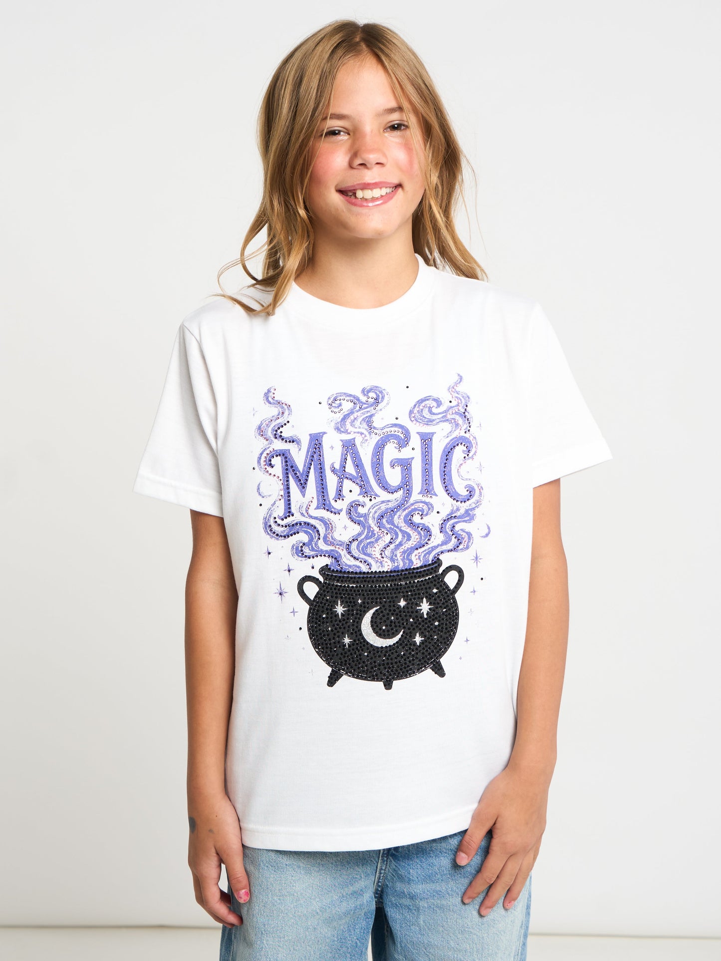A smiling young girl with blonde hair stands against a light gray studio background wearing a white graphic t-shirt with a purple and black cauldron design and the word 'MAGIC' in purple, paired with light blue jeans.