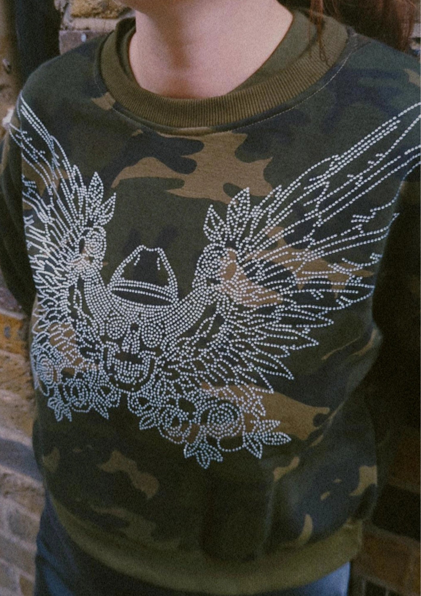Camouflage sweatshirt with rhinestone cowboy skull design worn by a  model against a brick wall.