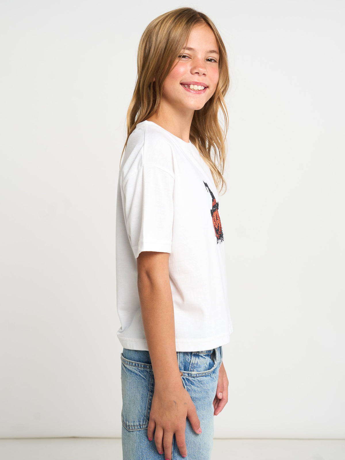 A young girl with blonde hair is shown from the side, wearing a white short-sleeve t-shirt with a small colorful graphic on the chest and light blue jeans, set against a plain white studio background.