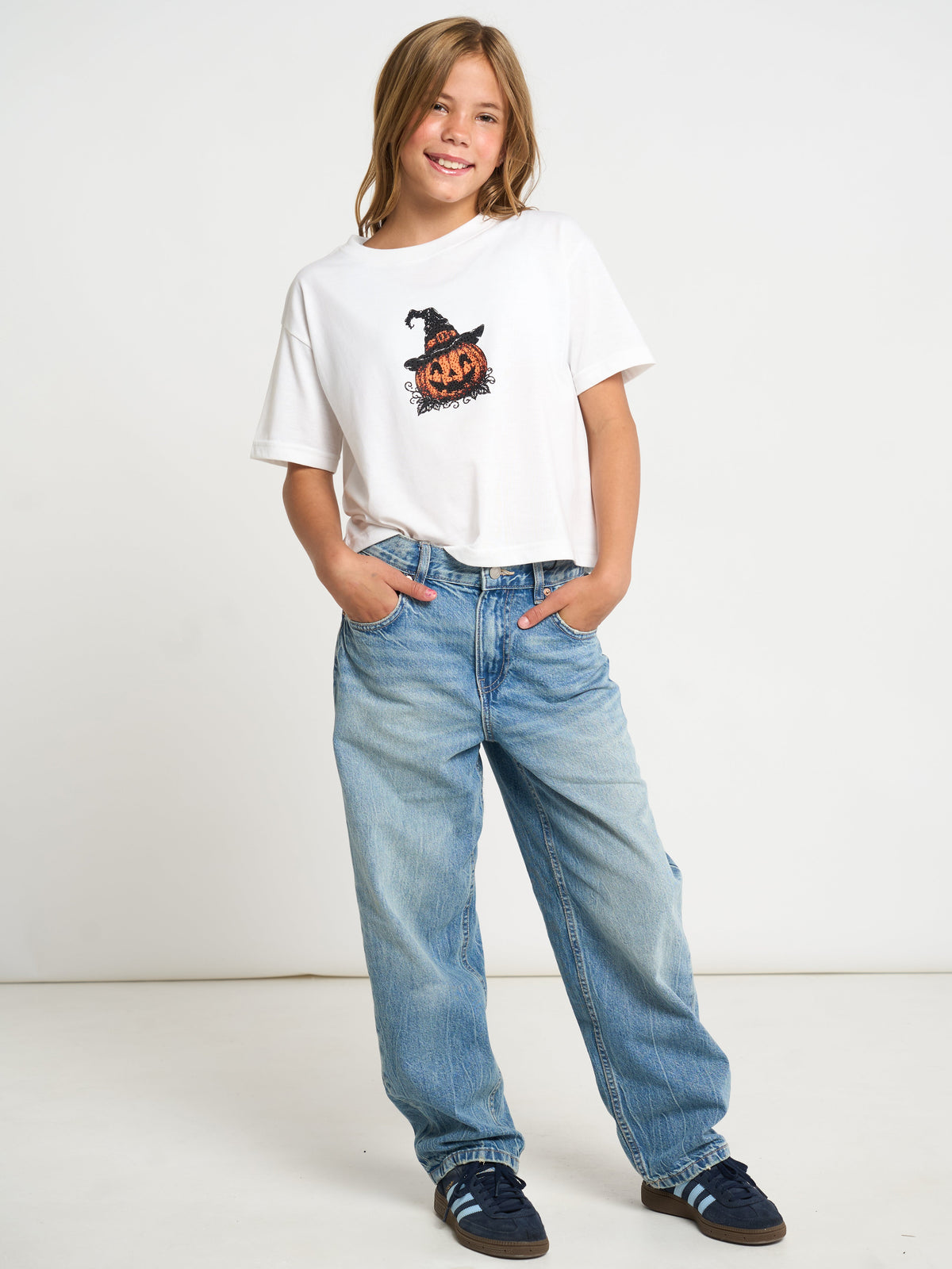 A child model wearing a white short-sleeve t-shirt with a black and orange pumpkin graphic, paired with light blue jeans and dark blue sneakers, standing against a plain white studio background.