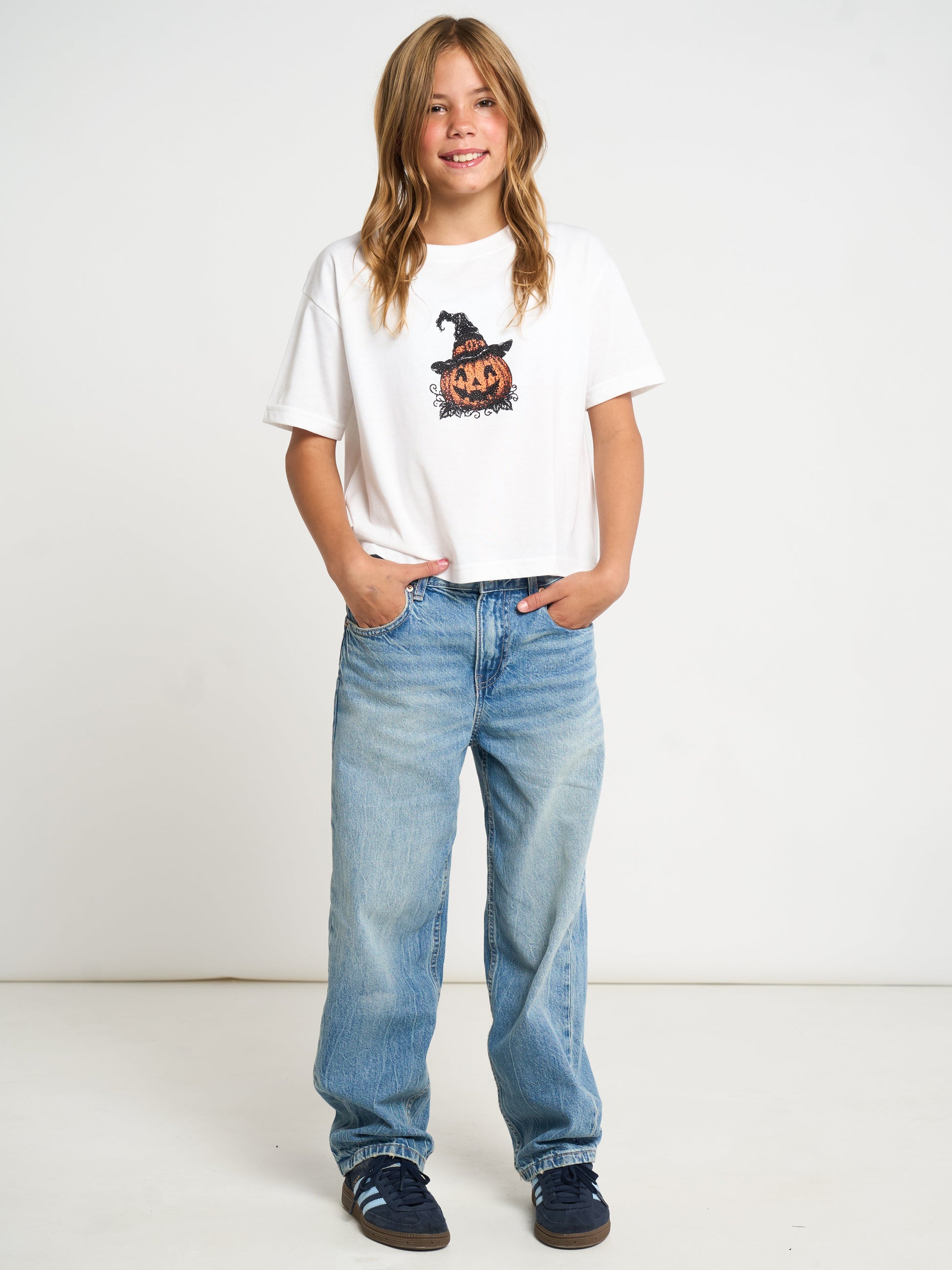 A young girl with blonde hair stands against a white studio background wearing a white graphic t-shirt with a pumpkin and witch hat design, light blue jeans, and dark blue sneakers.