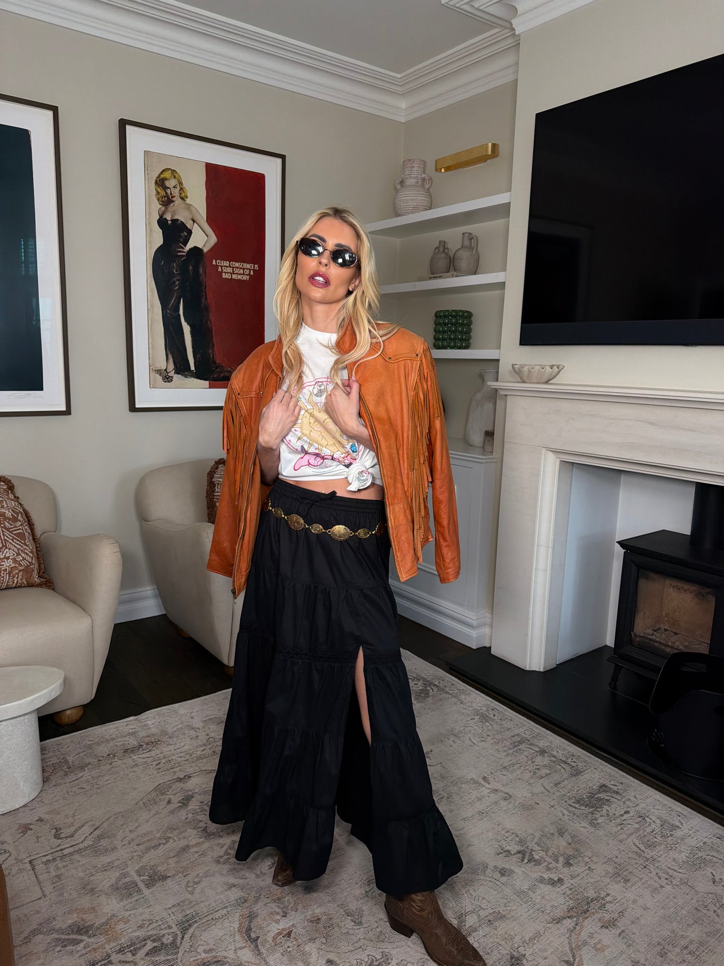 Woman wearing an oversized white graphic t-shirt featuring a celestial sun face design with butterfly and embellished details, styled with a black tiered skirt and statement belt in an indoor setting.