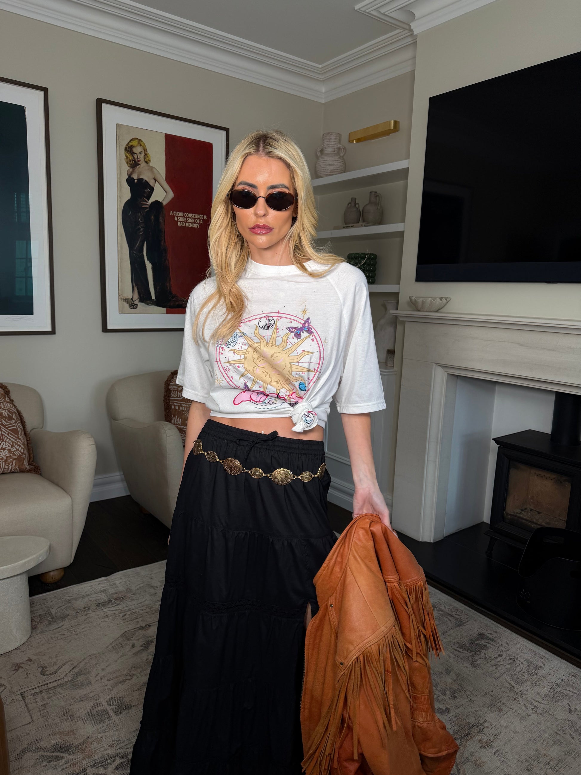 Woman in a living room wearing sunglasses and a white graphic t-shirt with a long black skirt.