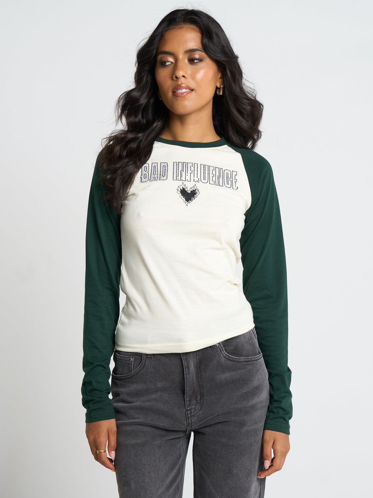 A woman modeling a long-sleeve raglan t-shirt with an off-white body and dark green sleeves. The shirt features the text 'BAD INFLUENCE' in a distressed arched font with a small black heart graphic below it.