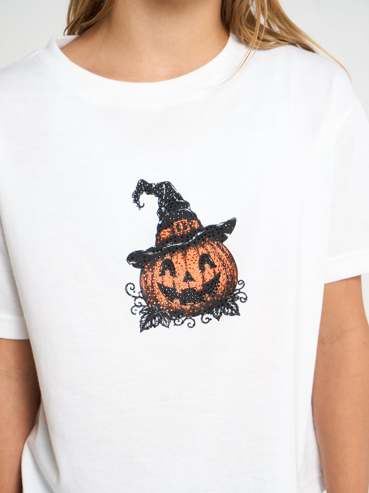 A white short-sleeve crewneck t-shirt featuring a black and orange Halloween graphic of a jack-o'-lantern wearing a witch's hat, worn by a model.