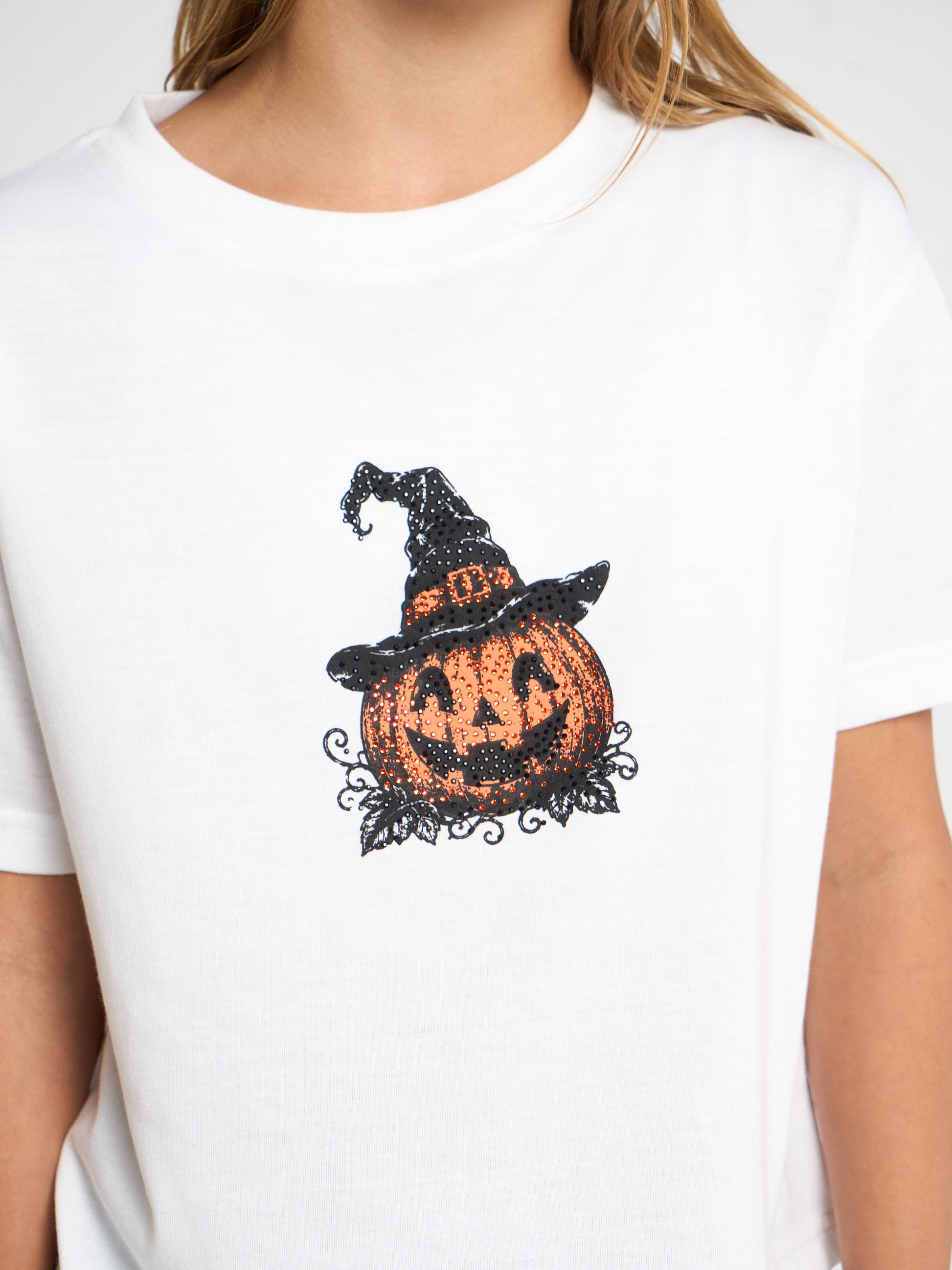 A white short-sleeve crewneck t-shirt featuring a black and orange Halloween graphic of a jack-o'-lantern wearing a witch's hat, worn by a model.