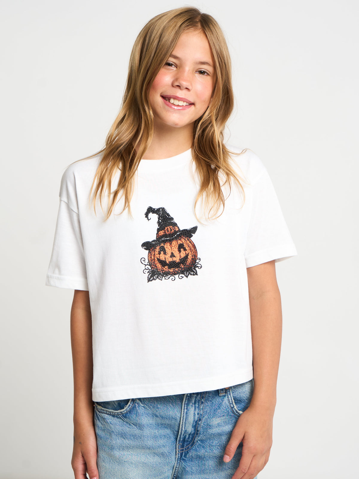 A young girl with blonde hair smiling while wearing a white short-sleeve t-shirt with a graphic of a jack-o'-lantern pumpkin wearing a witch hat, paired with blue jeans, against a light gray studio background.