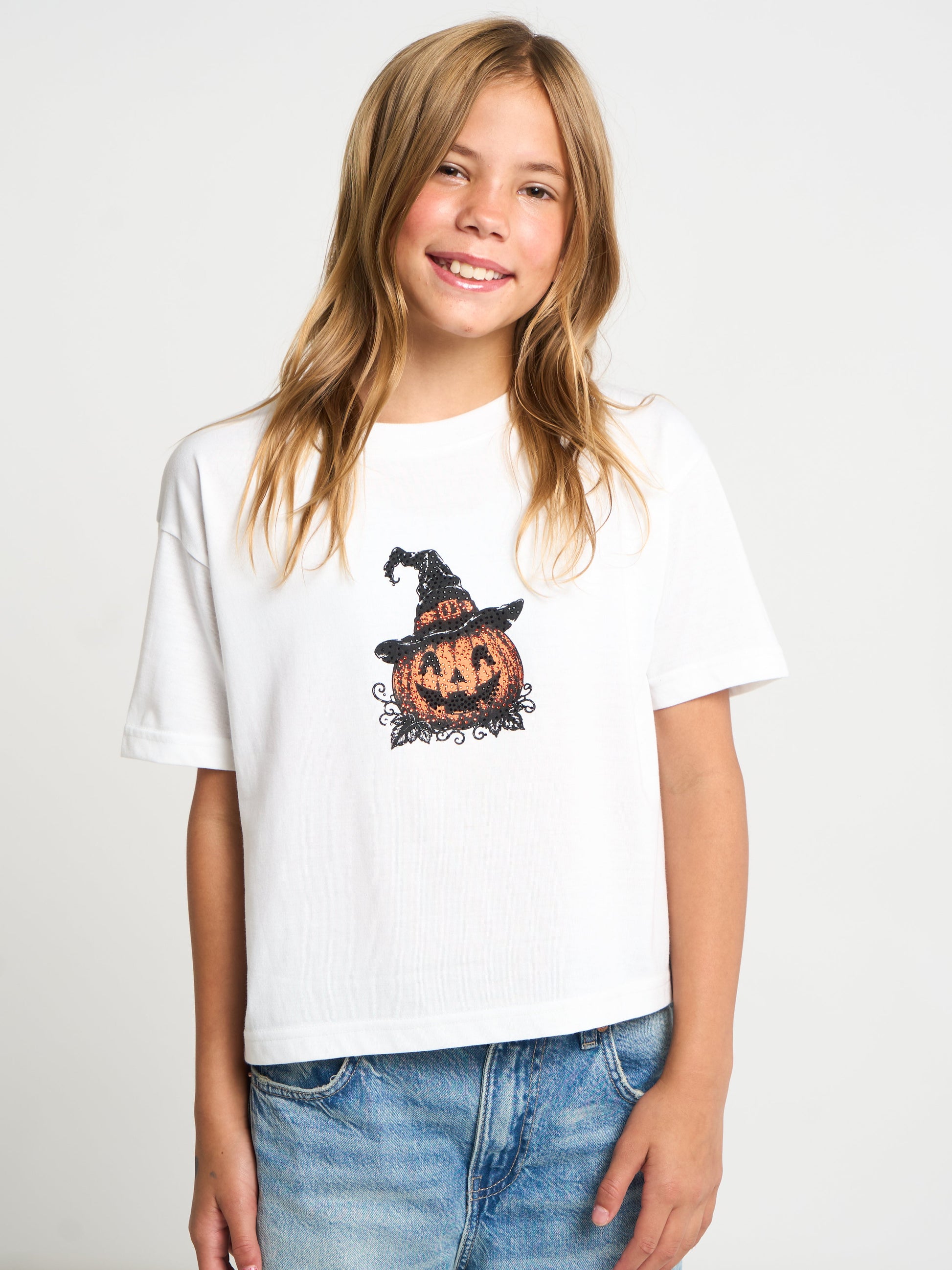 A young girl with blonde hair smiling while wearing a white short-sleeve t-shirt with a graphic of a jack-o'-lantern pumpkin wearing a witch hat, paired with blue jeans, against a light gray studio background.