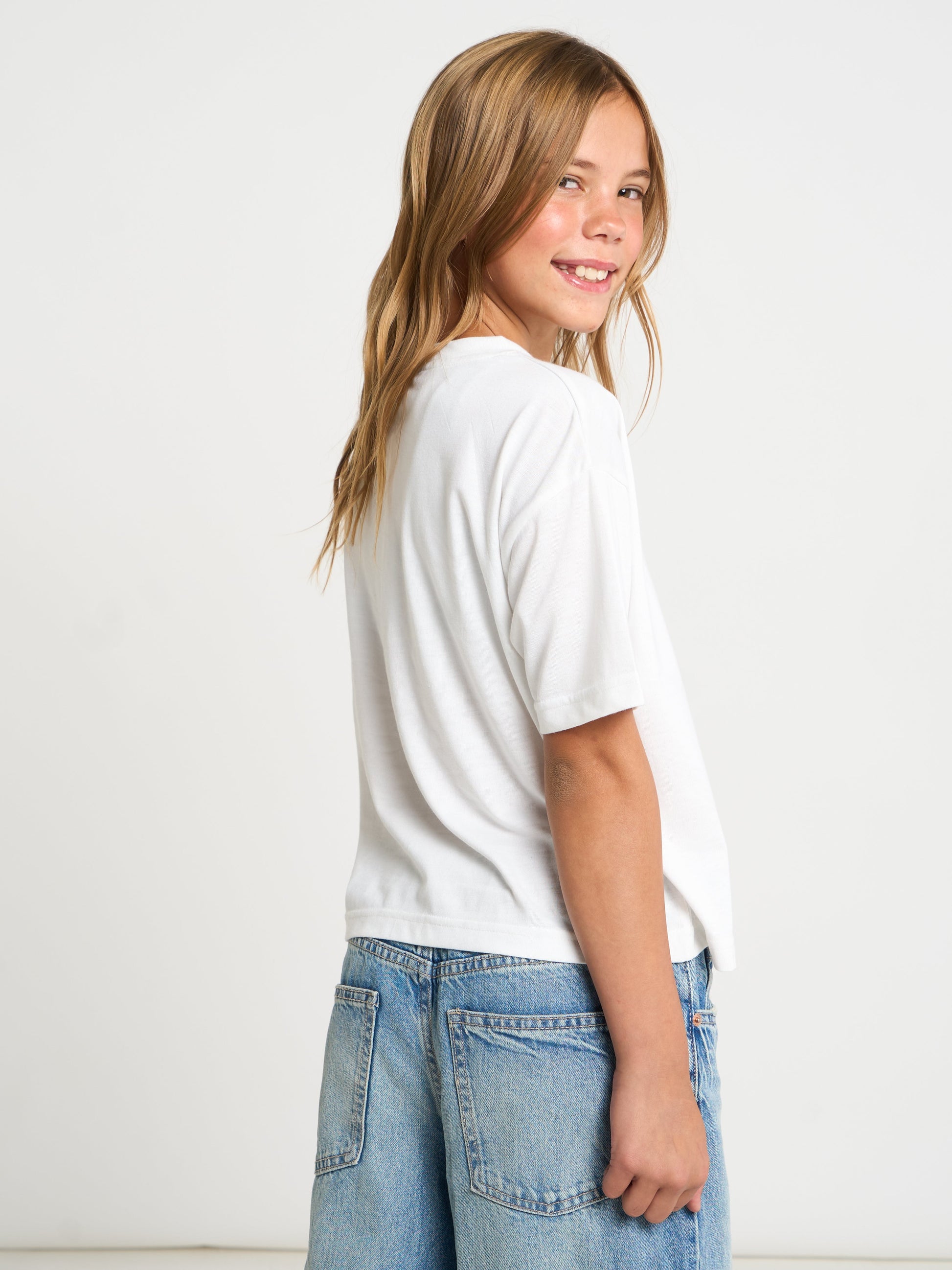A young girl with long blonde hair is shown from a side-back angle, wearing a plain white short-sleeve t-shirt and light blue denim jeans against a clean white studio background.