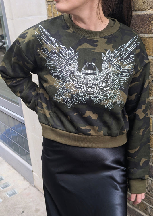 Person wearing a camouflage sweatshirt with a rhinestone skull design on a brick wall background