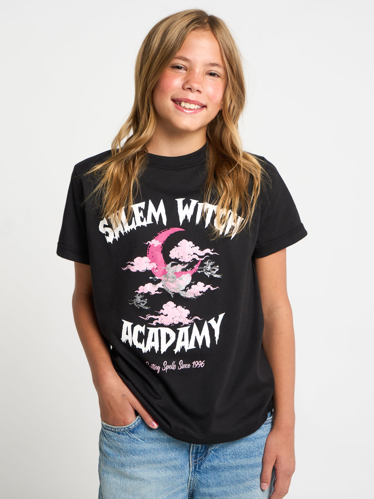 A young girl with blonde hair smiling and wearing a black graphic t-shirt that reads 'Salem Witch Academy' with a pink and white bird design, paired with blue jeans, against a plain white background.