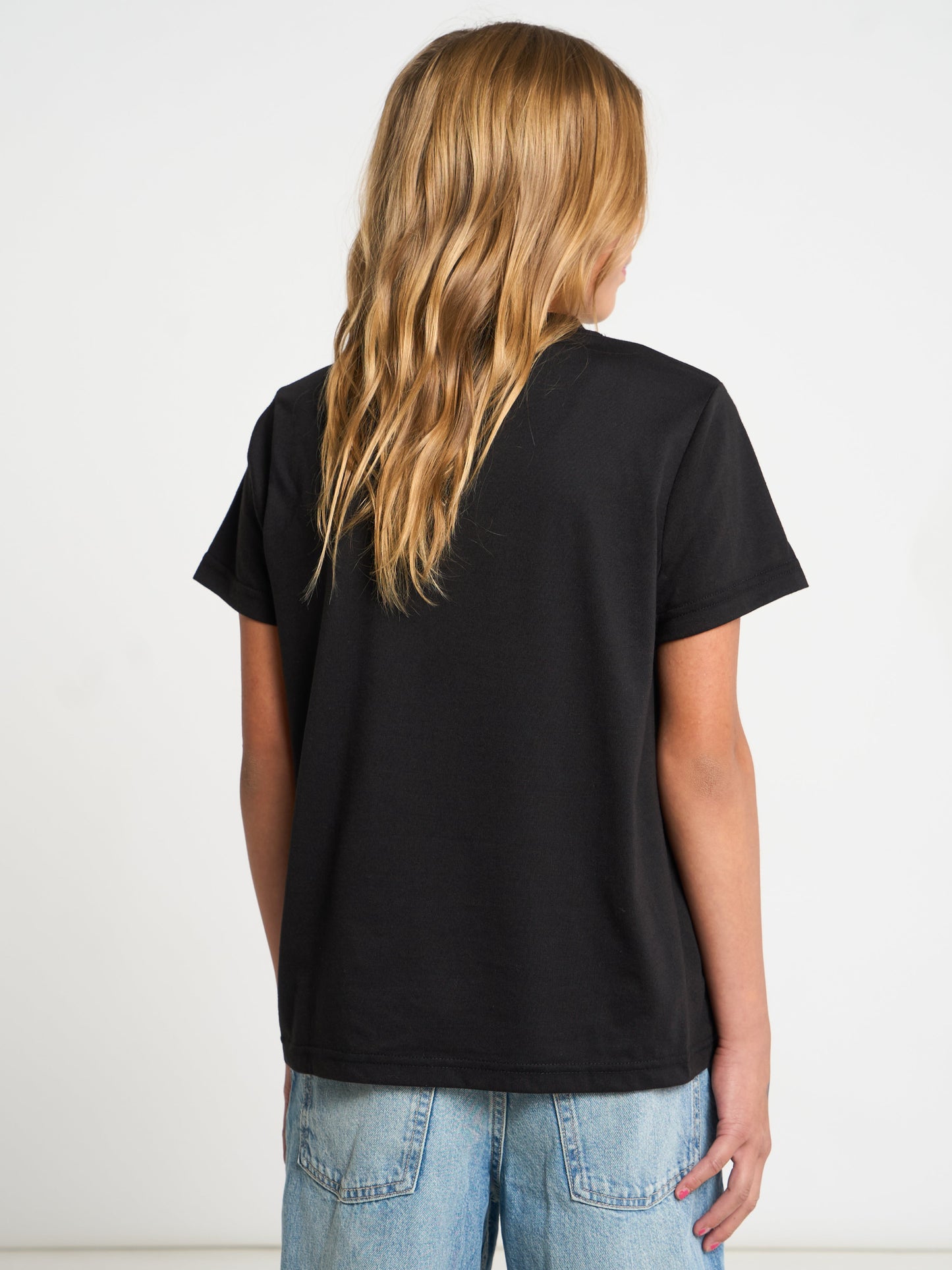 A girl with long blonde hair is shown from behind wearing a black short-sleeve t-shirt and light blue jeans against a plain white background.