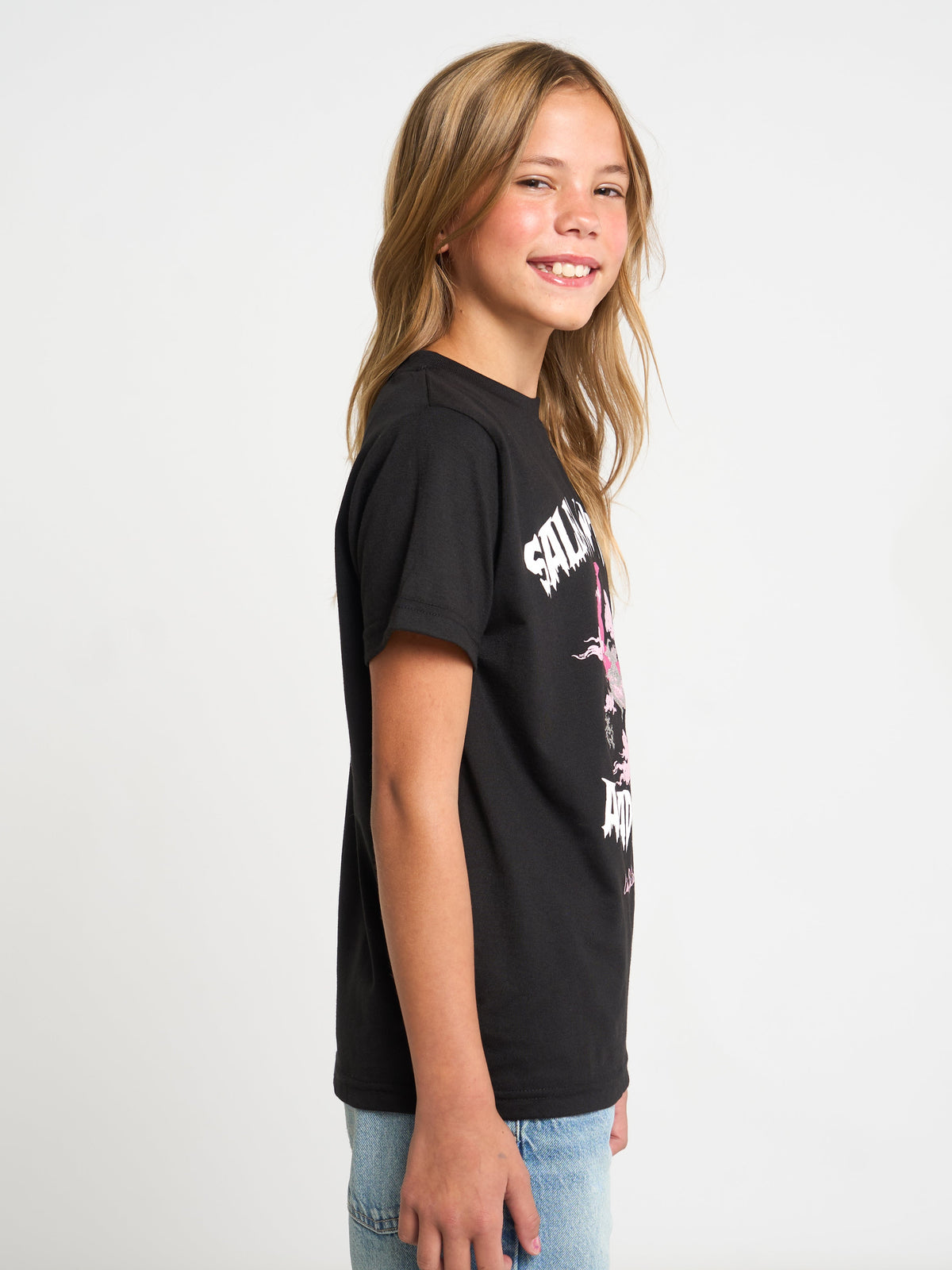 A young girl with blonde hair is shown from the side profile, wearing a black short-sleeve graphic t-shirt with white and pink text, paired with light blue jeans, against a plain white studio background.