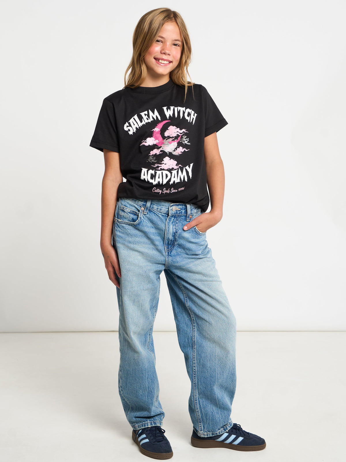 A young girl modeling a black graphic t-shirt that reads 'Salem Witch Academy' with a pink witch illustration, paired with light-wash blue jeans and dark blue sneakers, against a plain white studio background.