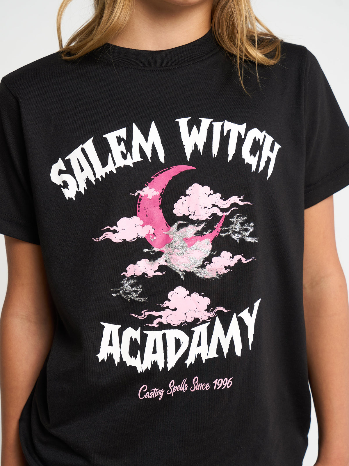 A person wearing a black graphic T-shirt with the words 'SALEM WITCH ACADEMY' in white distressed lettering and a pink crescent moon/cloud design on the front.