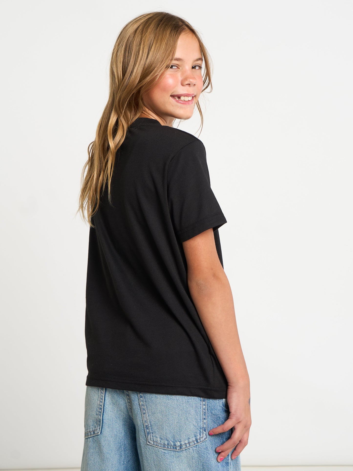 A young girl with blonde hair is shown from a back-three-quarter angle, wearing a black short-sleeve t-shirt and light blue denim jeans against a plain white studio background.