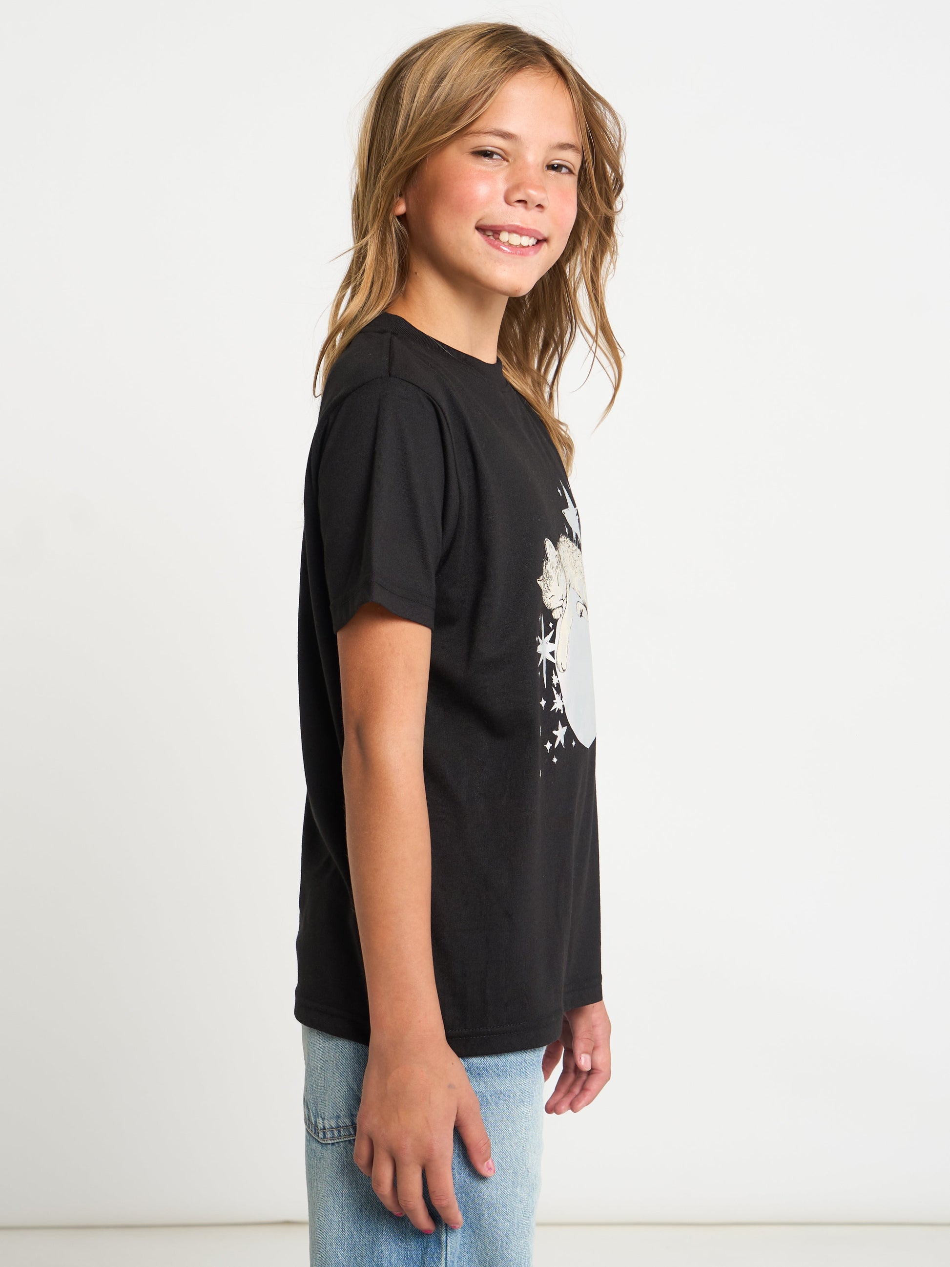 A young girl with blonde hair is shown from the side, wearing a black short-sleeve t-shirt with a white graphic print on the front and light blue jeans, set against a plain white studio background.