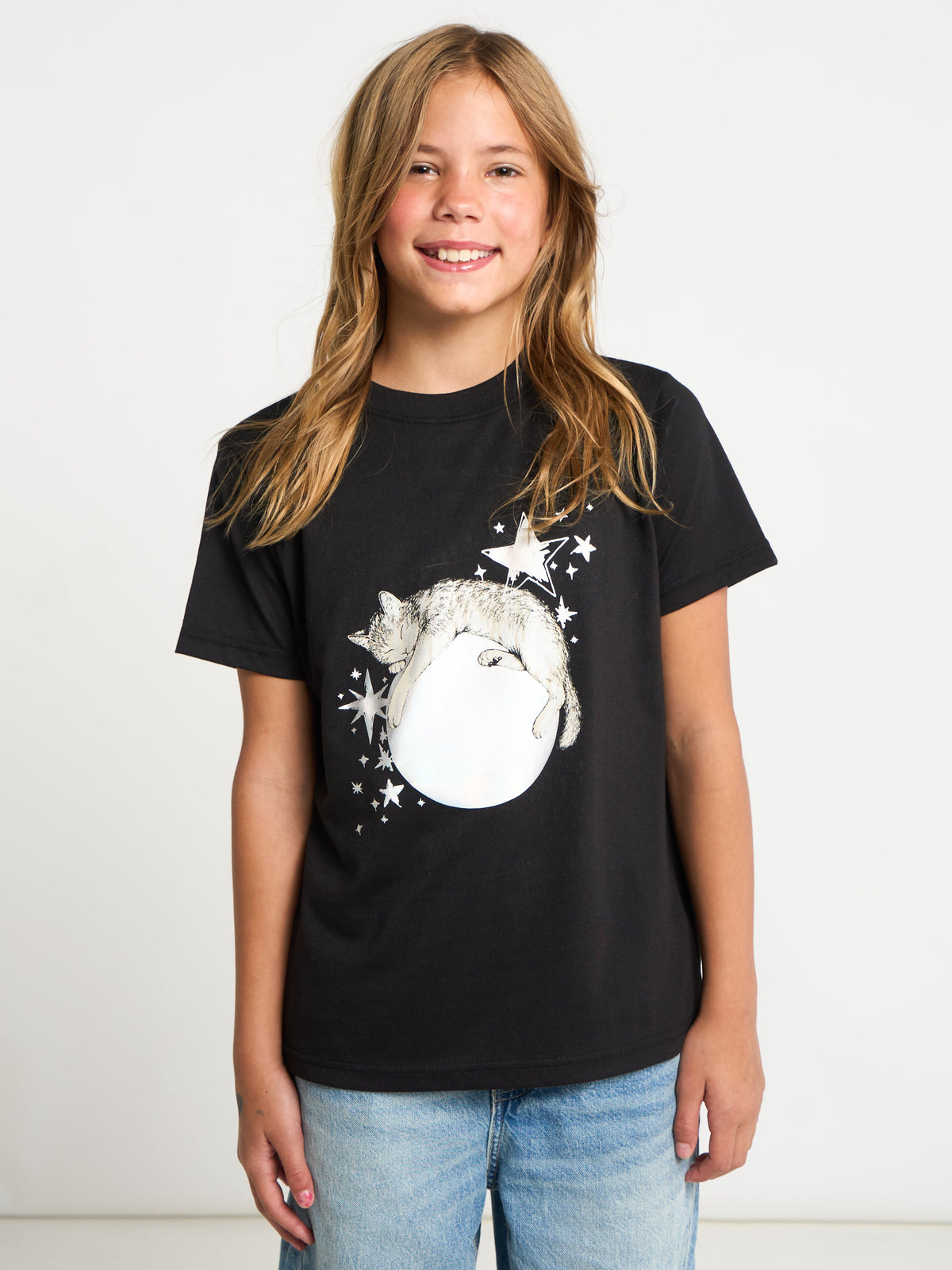 A young girl with long blonde hair smiling, wearing a black short-sleeve t-shirt with a white graphic of a cat on a crescent moon surrounded by stars, paired with light blue jeans, against a plain white background.