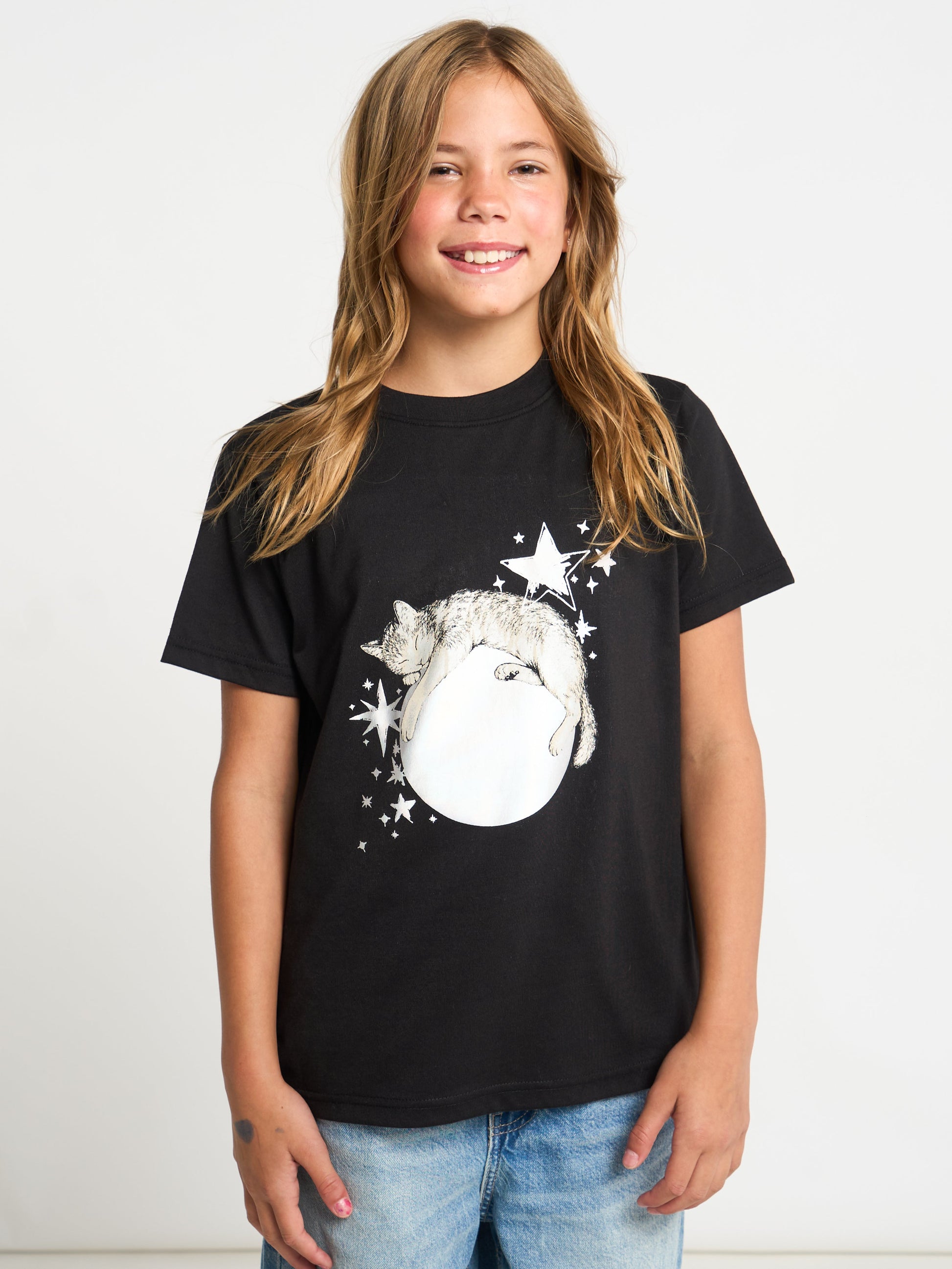 A young girl with long light brown hair smiling, wearing a black short-sleeve graphic t-shirt with a white cat and moon design, paired with blue jeans, against a plain white background.