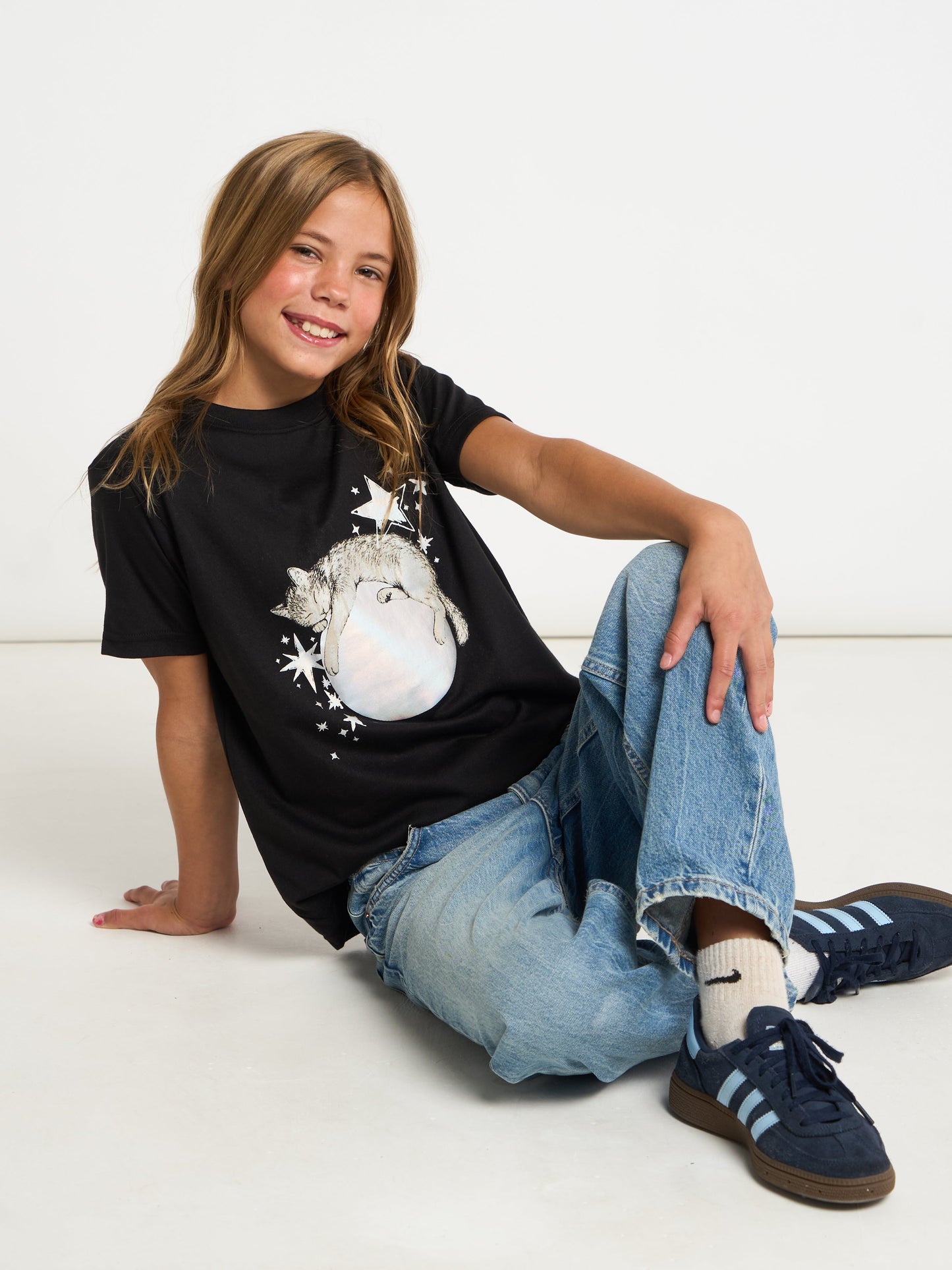 A young girl with blonde hair sits on a white floor wearing a black graphic t-shirt with a white snowman design, blue jeans, white socks, and navy blue sneakers.