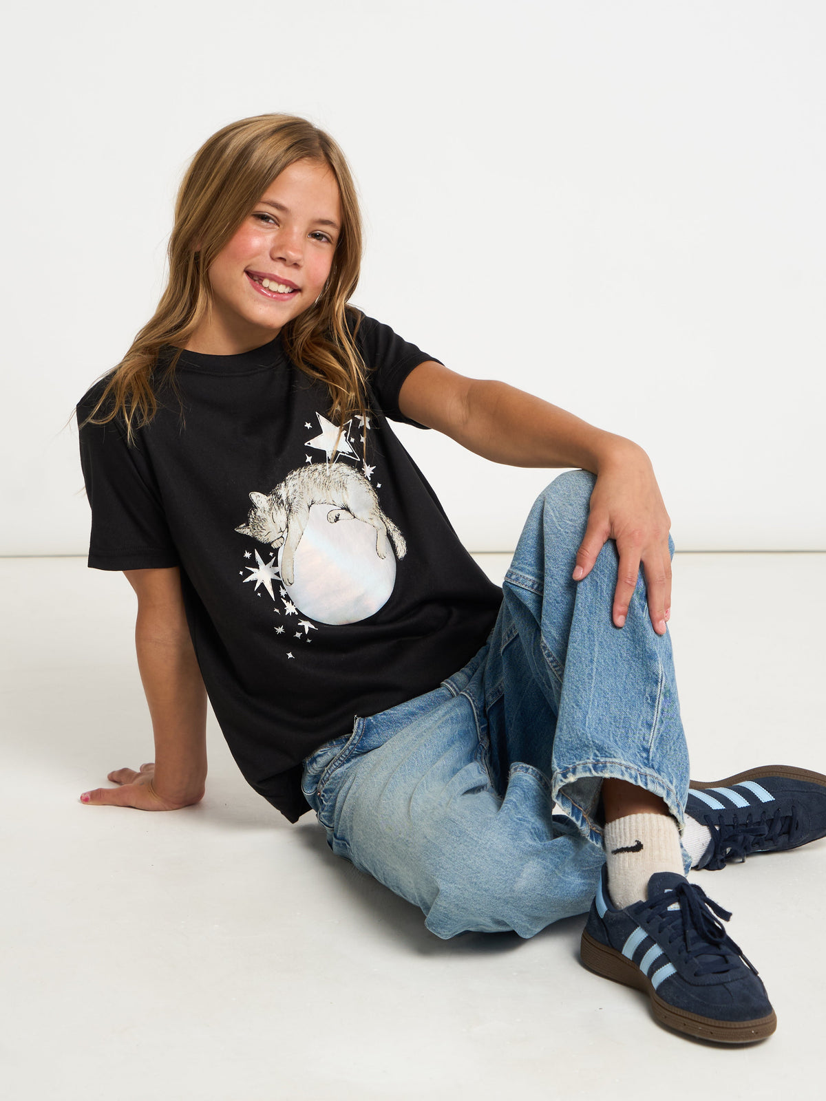 A young girl with blonde hair sits on a white floor wearing a black graphic t-shirt with a white snowman design, blue jeans, white socks, and navy blue sneakers.