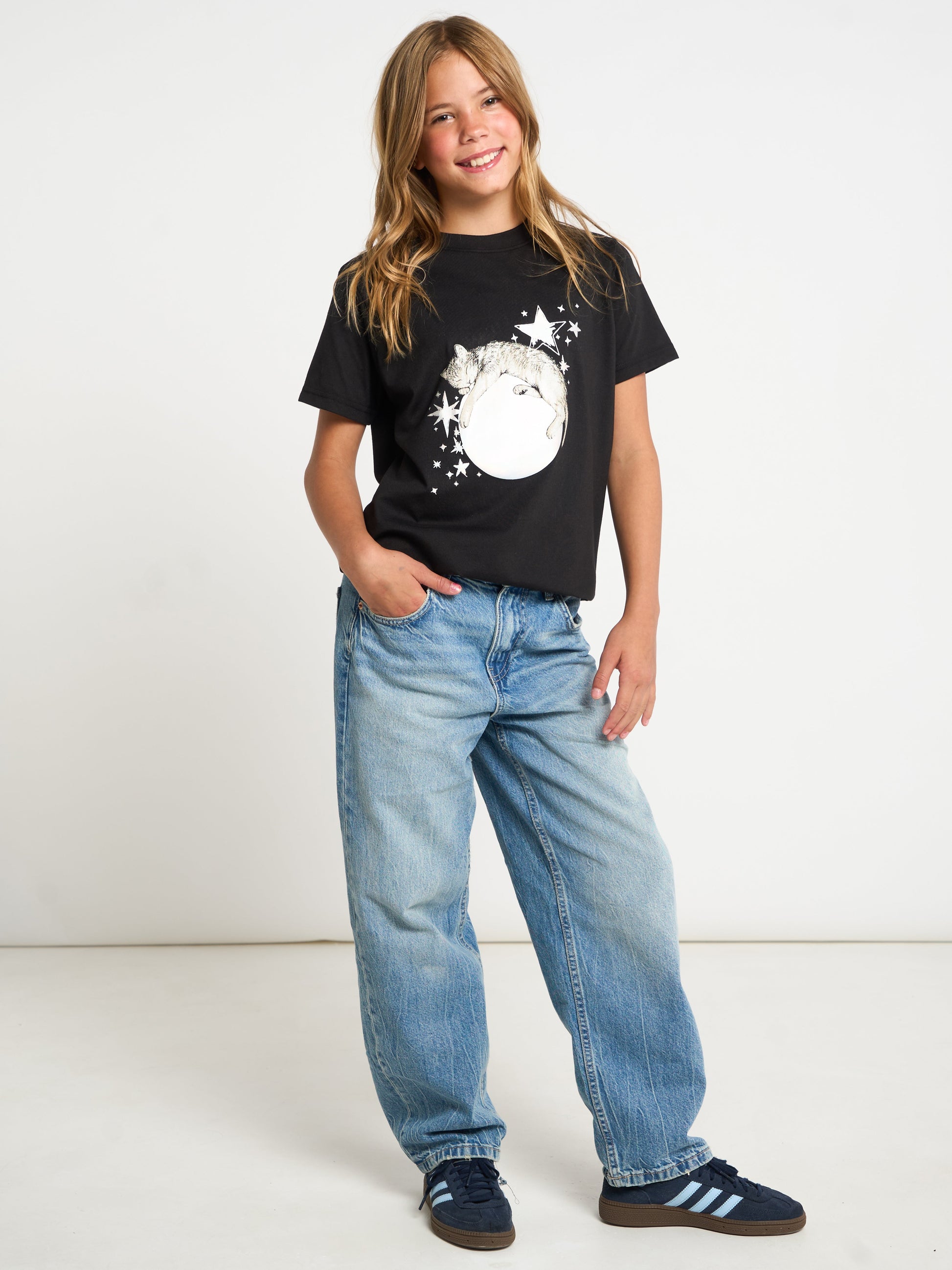 A young girl with long blonde hair stands smiling, wearing a black graphic t-shirt with a white cat and moon design, light blue jeans, and navy blue sneakers with white stripes, set against a plain white studio background.