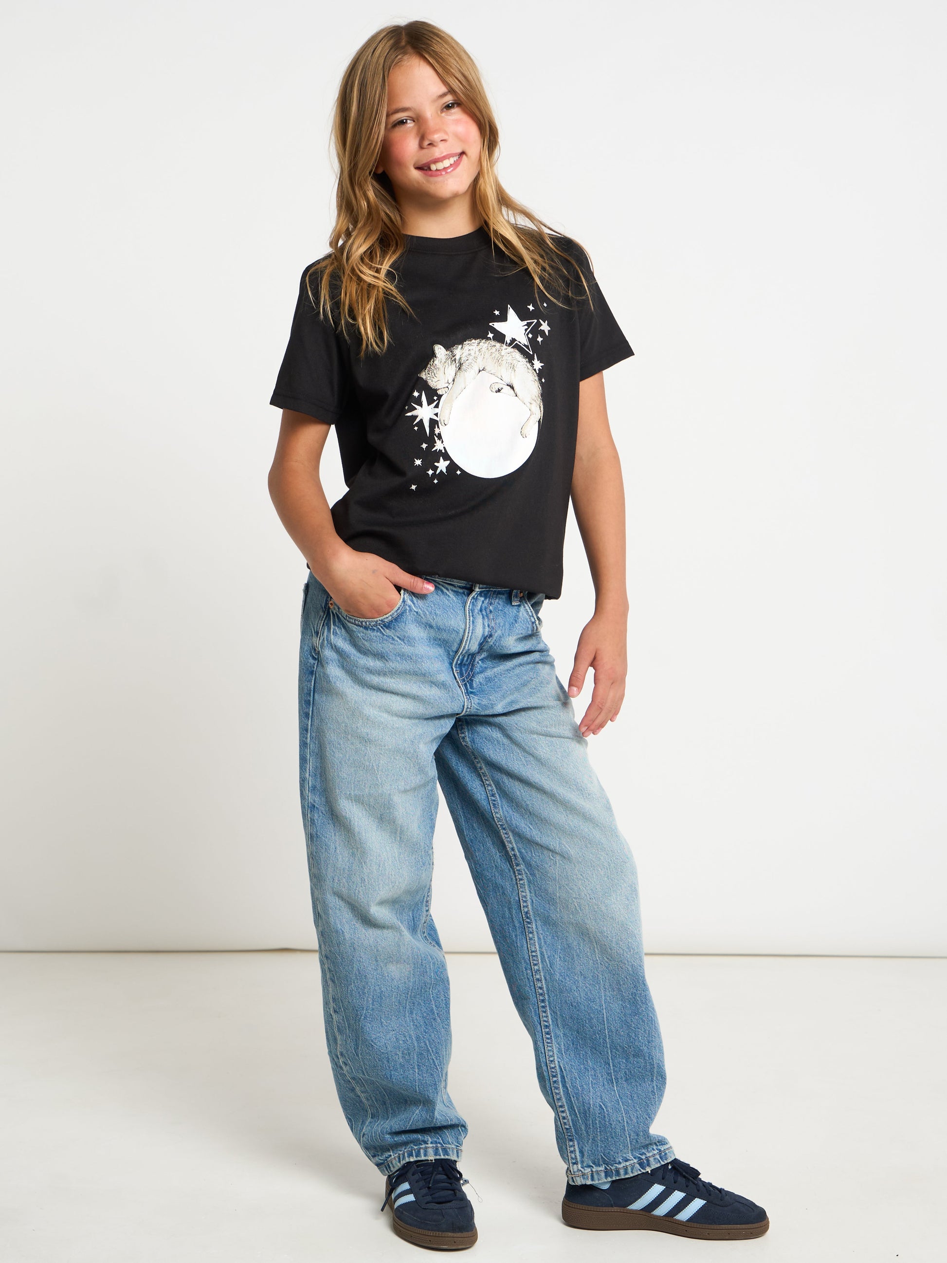 A young girl modeling a black graphic t-shirt with a white moon design, paired with light wash blue jeans and navy blue sneakers, standing against a plain white studio background.