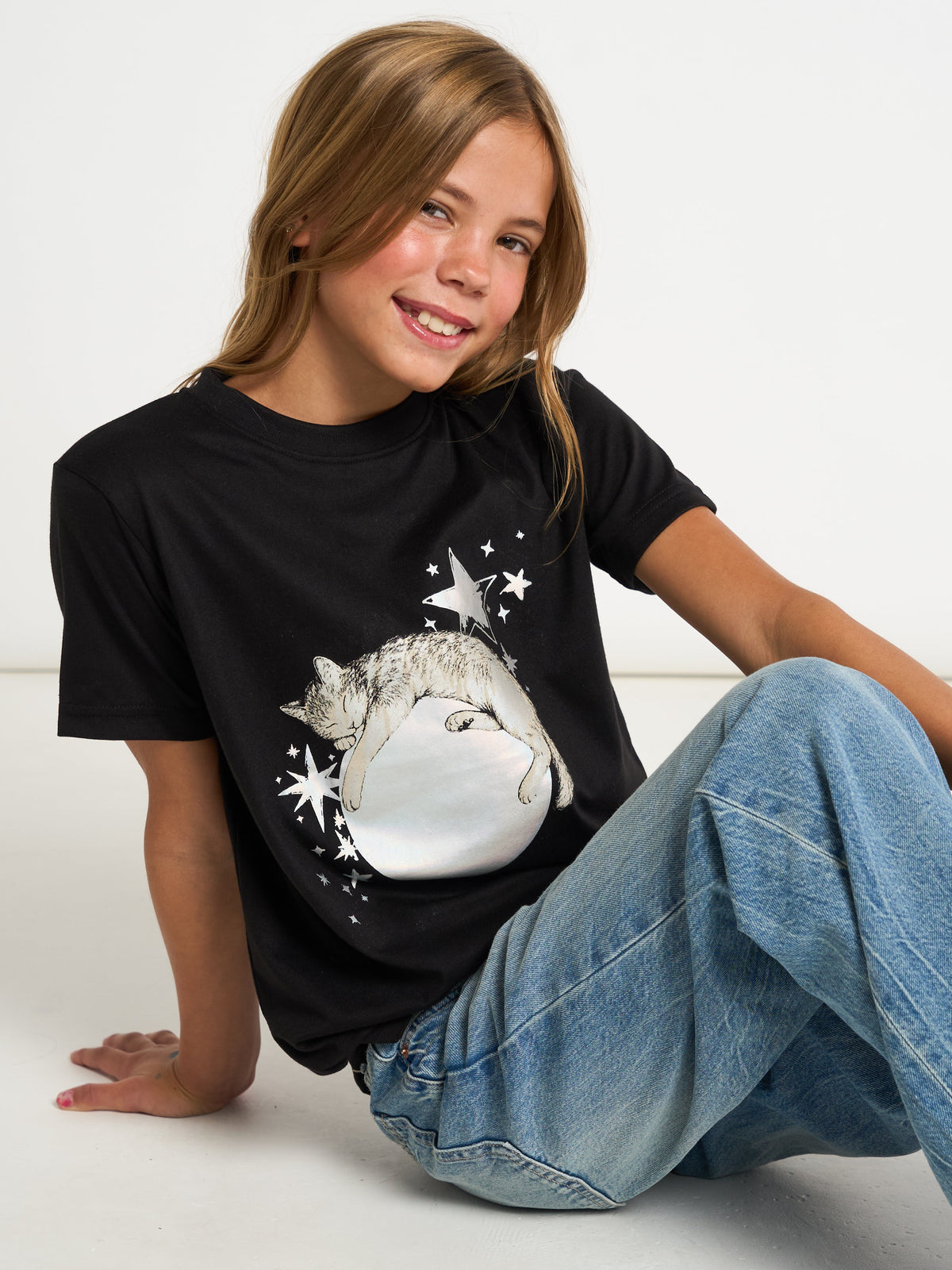 A young girl with blonde hair sits on a white floor wearing a black graphic t-shirt with a sleeping cat design and blue jeans.