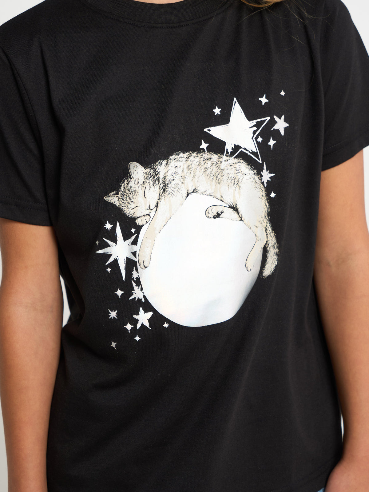 A person wearing a black short-sleeve T-shirt with a large graphic print of a sleeping cat curled up inside a white egg, surrounded by white stars.