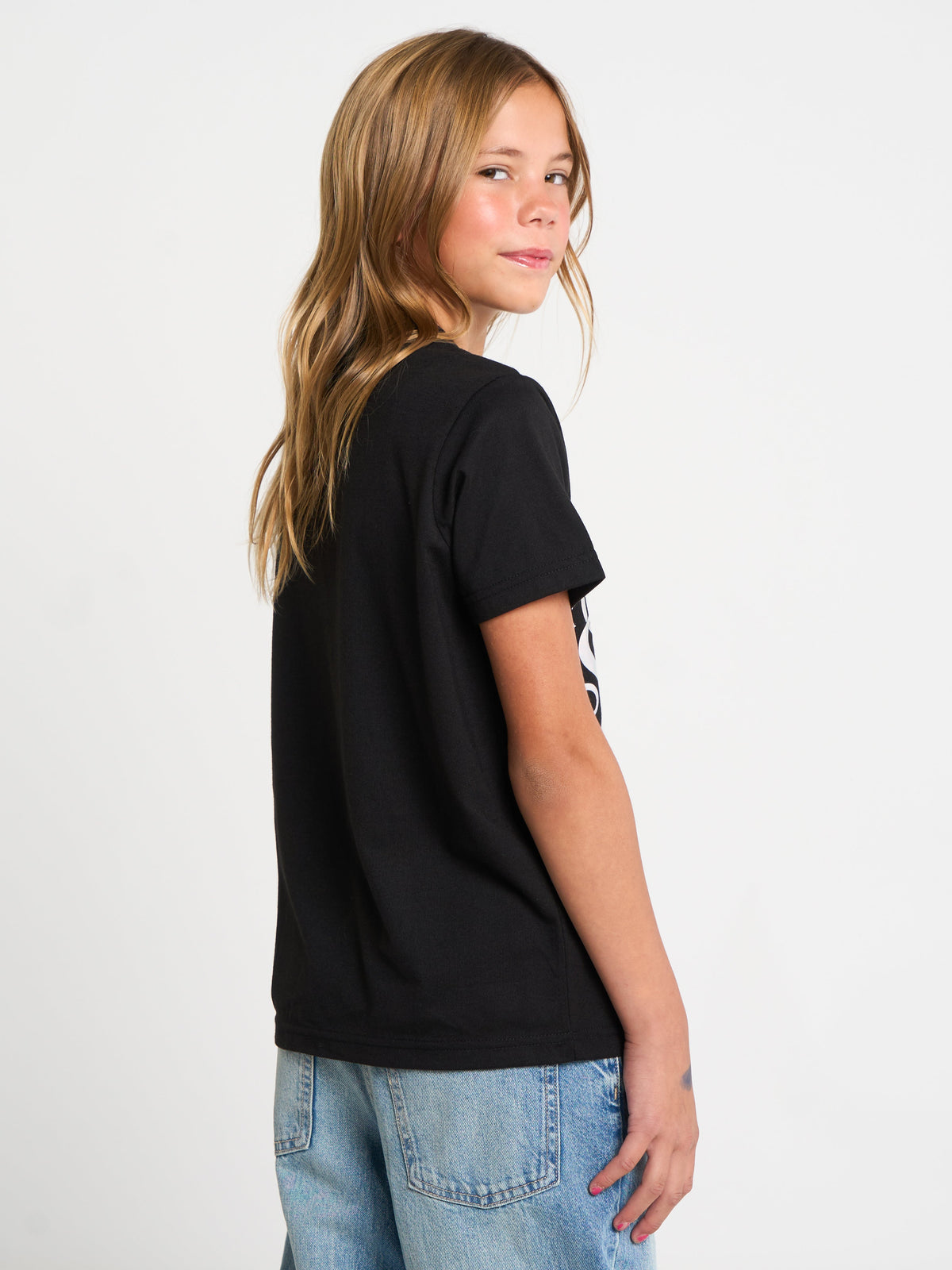 A young girl with long light brown hair is shown from a side-back angle wearing a black short-sleeve t-shirt and light blue denim jeans against a plain white studio background.