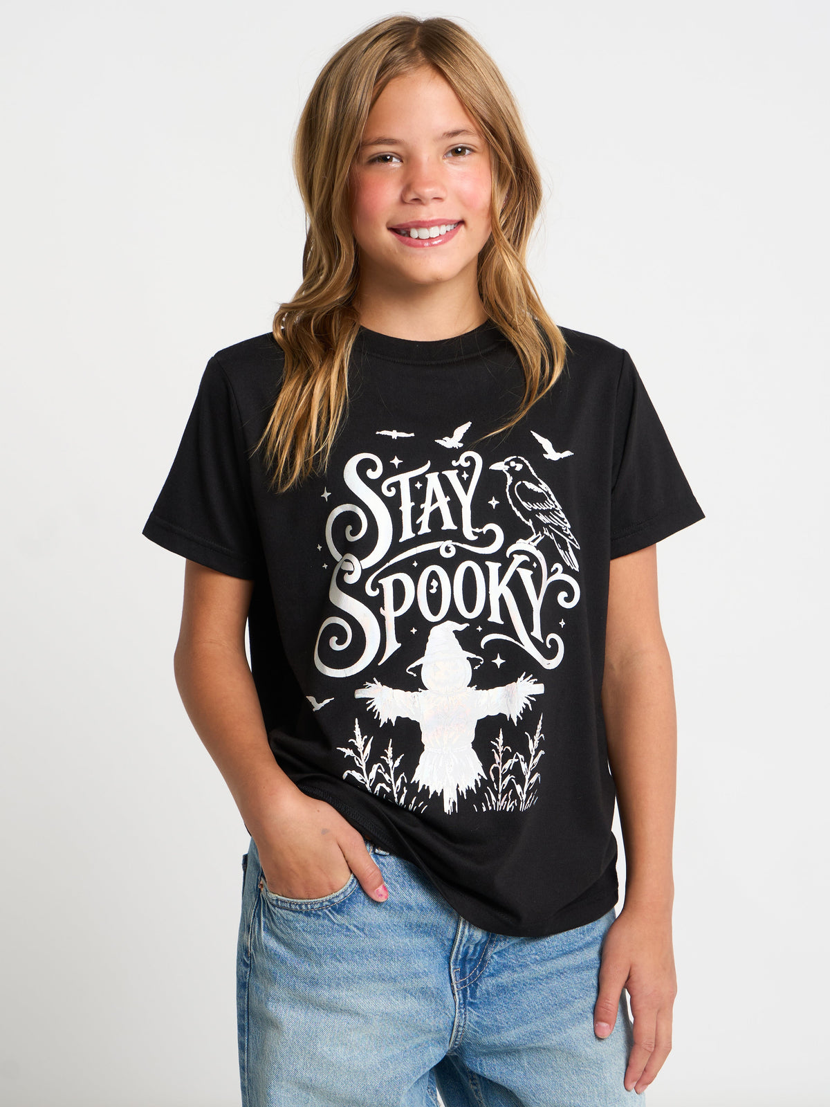 A young girl with blonde hair smiling and wearing a black graphic t-shirt with a white 'Stay Spooktacular' scarecrow design, paired with blue jeans, against a plain white background.