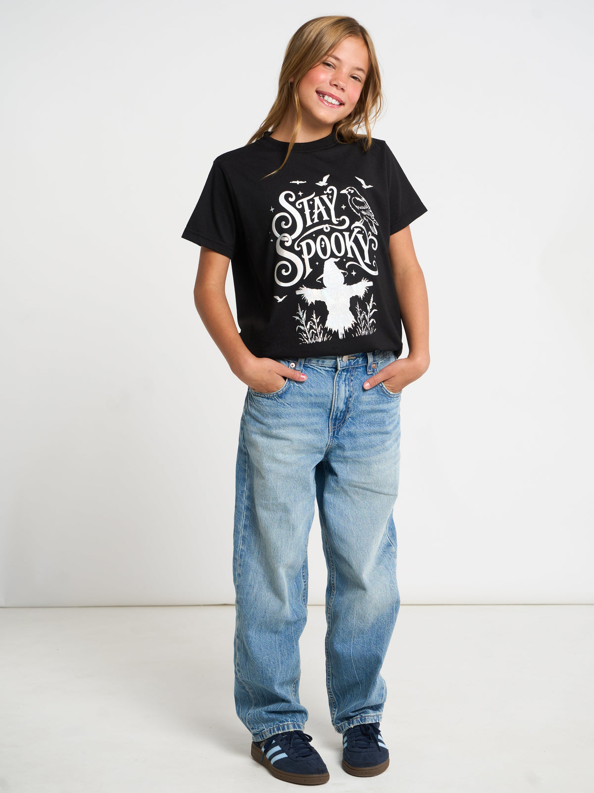 A young girl smiling and posing in a black graphic t-shirt with 'Stay Spooktacular' printed in white, paired with light-wash blue jeans and dark blue sneakers, set against a plain white studio background.