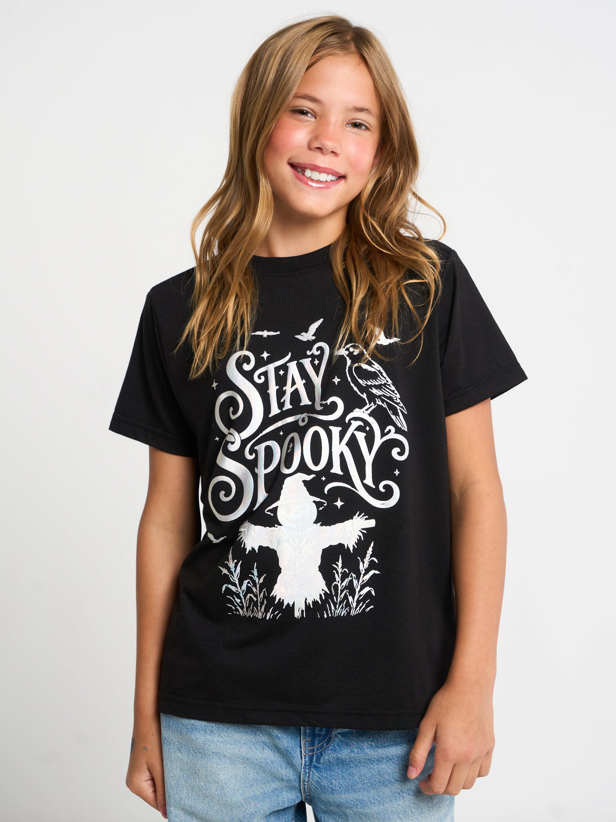 A young girl with long light brown hair smiling, wearing a black short-sleeve graphic t-shirt with a white 'Stay Spooky' design featuring a scarecrow, crows, and bats, paired with blue jeans against a light gray studio background.